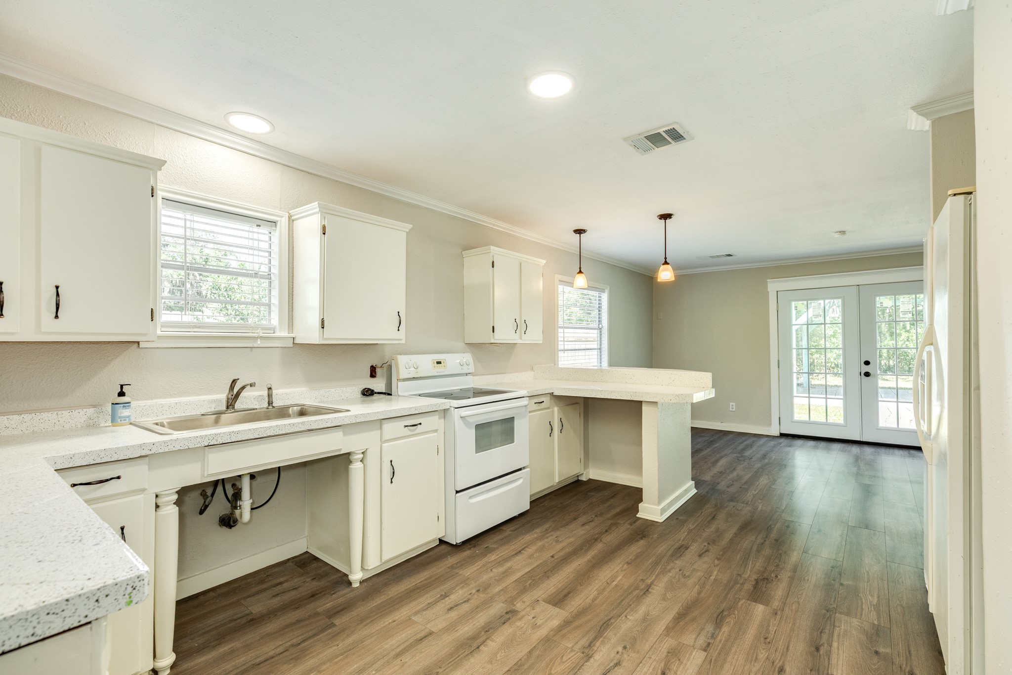 Spacious kitchen features white cabinetry, speckled countertops, a double-basin sink, and a breakfast bar overlooking the dining area. Wood-look flooring, pendant lighting, and French doors create an inviting space for everyday living and entertaining.