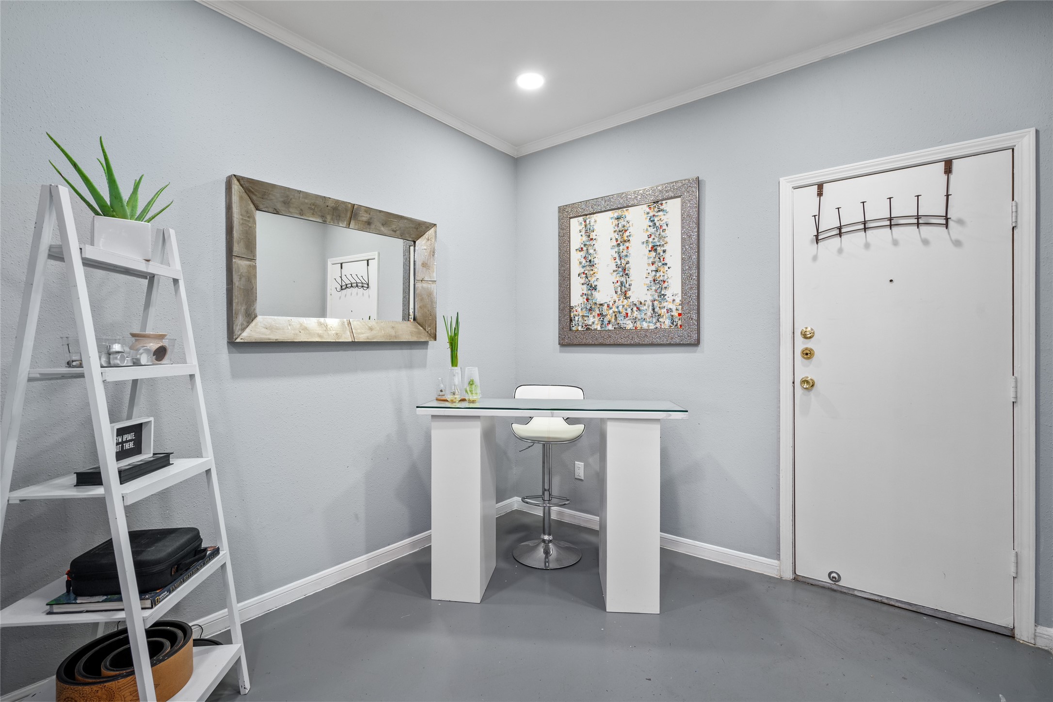 This inviting entryway doubles as a sleek and modern workspace. Love how the owners designed this area featuring a glass-top desk, a contemporary white barstool, and a decorative ladder shelf for stylish storage. Soft gray walls complement the rustic wood-framed mirror and abstract artwork, creating a welcoming and functional space.