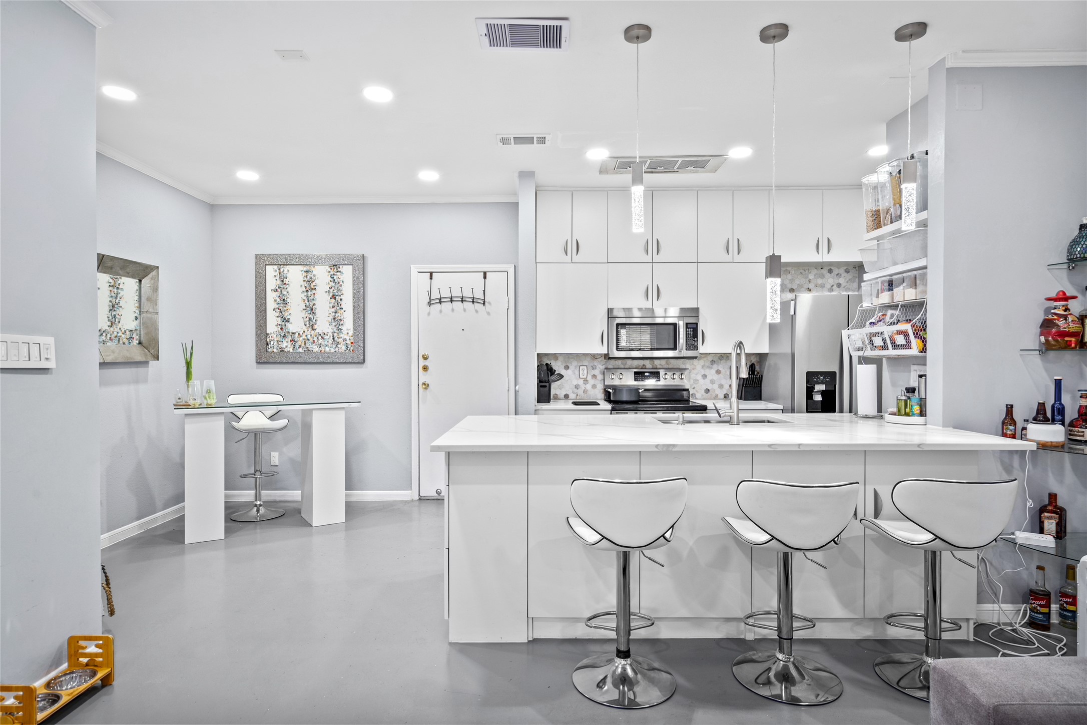 This stylish kitchen features crisp white cabinetry, stainless steel appliances, and a spacious island with a waterfall countertop, perfect for casual dining or entertaining. Elegant pendant lighting and a hexagonal tile backsplash add a contemporary touch, while open shelving provides functional storage. The seamless flow into the living area makes this kitchen both inviting and practical.