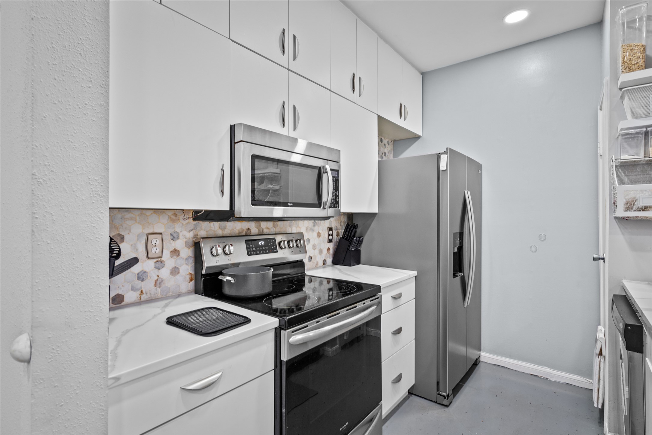 This compact yet highly functional kitchen showcases crisp white cabinetry, a stylish hexagonal backsplash, and quartz countertops. Stainless steel appliancesincluding a microwave, electric range, and spacious refrigeratorprovide modern convenience, while ample overhead cabinets maximize storage. The clean, streamlined design makes meal prep and cooking a breeze.