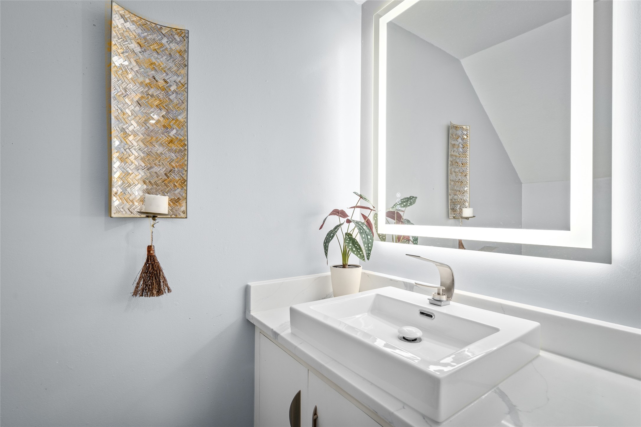 This elegant downstairs half bath features a modern vessel sink atop a sleek quartz countertop. A backlit mirror adds a contemporary touch, while the neutral tones and decorative wall accents create a warm and inviting space. Perfect for guests, this thoughtfully designed powder room combines function and style.