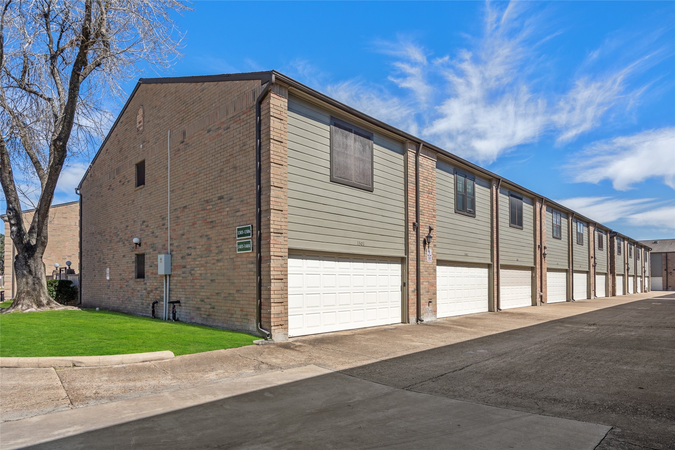 Rear view of the well-maintained townhome community, showcasing a row of two-car garages with clean white doors. The brick and siding exterior provides a classic and modern blend, while upper-level windows invite natural light inside. The paved driveway offers ample space for parking and easy access. A neatly landscaped green space with a mature tree adds a touch of nature to the area.