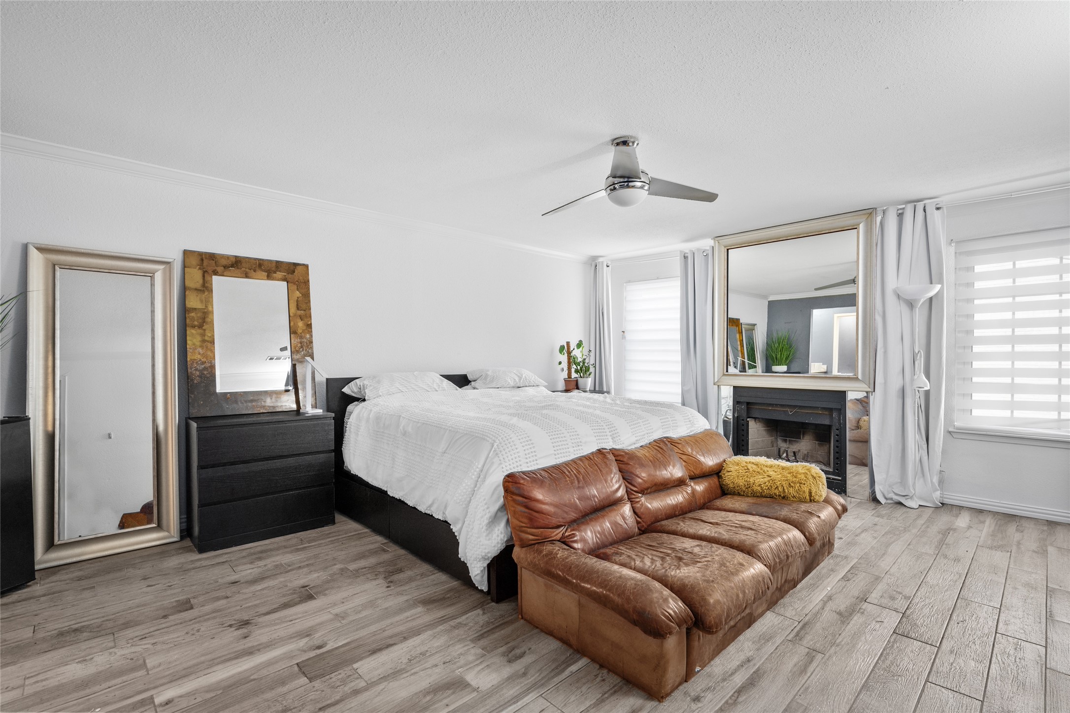 This expansive primary bedroom offers a serene retreat with its neutral tones, wood-look flooring, and abundant natural light. The cozy fireplace, framed by a statement mirror, adds warmth and character, while the modern ceiling fan ensures year-round comfort. Dual windows with sleek blinds provide adjustable privacy and brightness