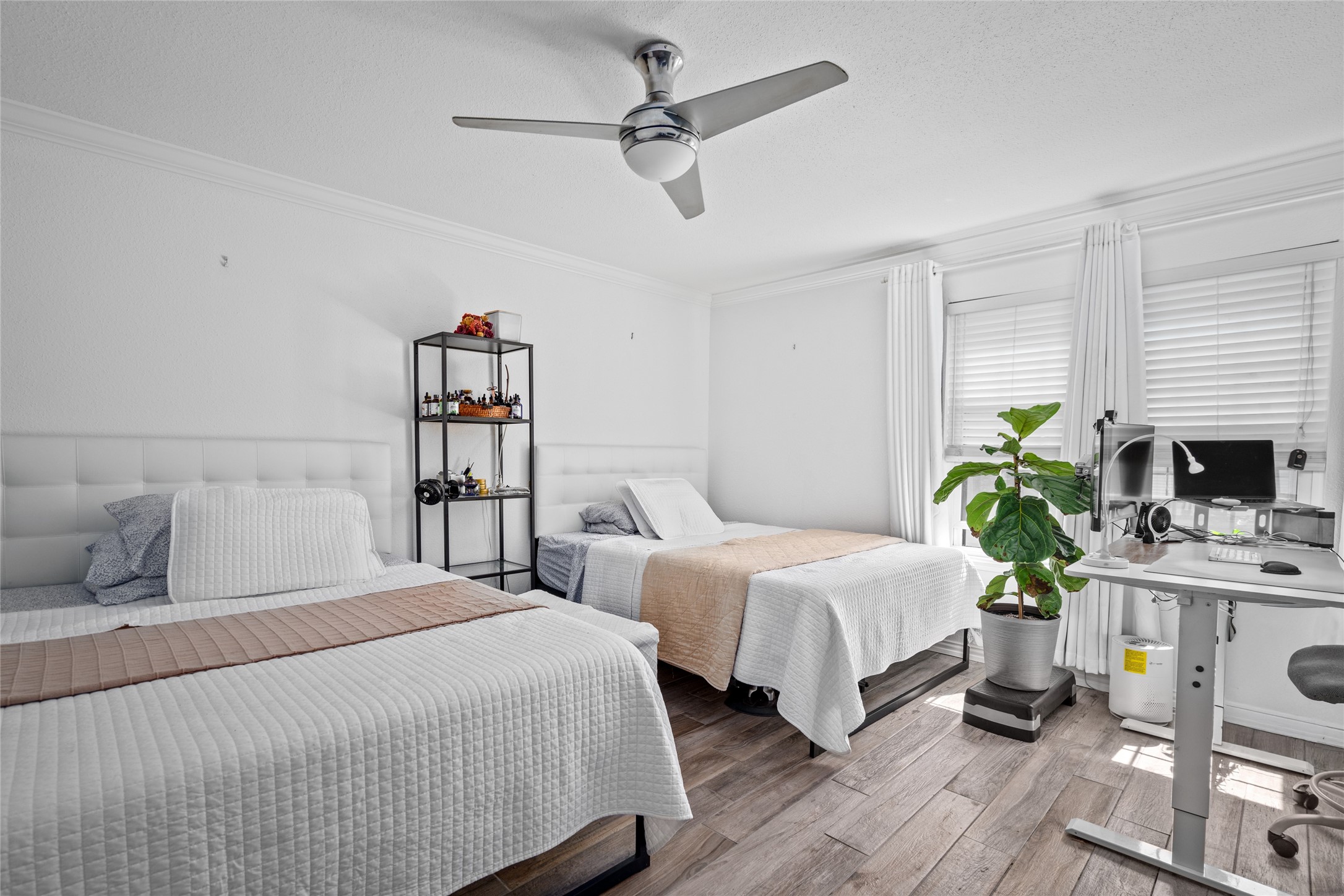 This well-lit secondary bedroom features ample space, large windows with elegant coverings, and a neutral color palette that enhances its bright and airy feel. The wood-look flooring adds warmth and style, while the ceiling fan provides comfort. A versatile layout makes this room ideal for various uses.