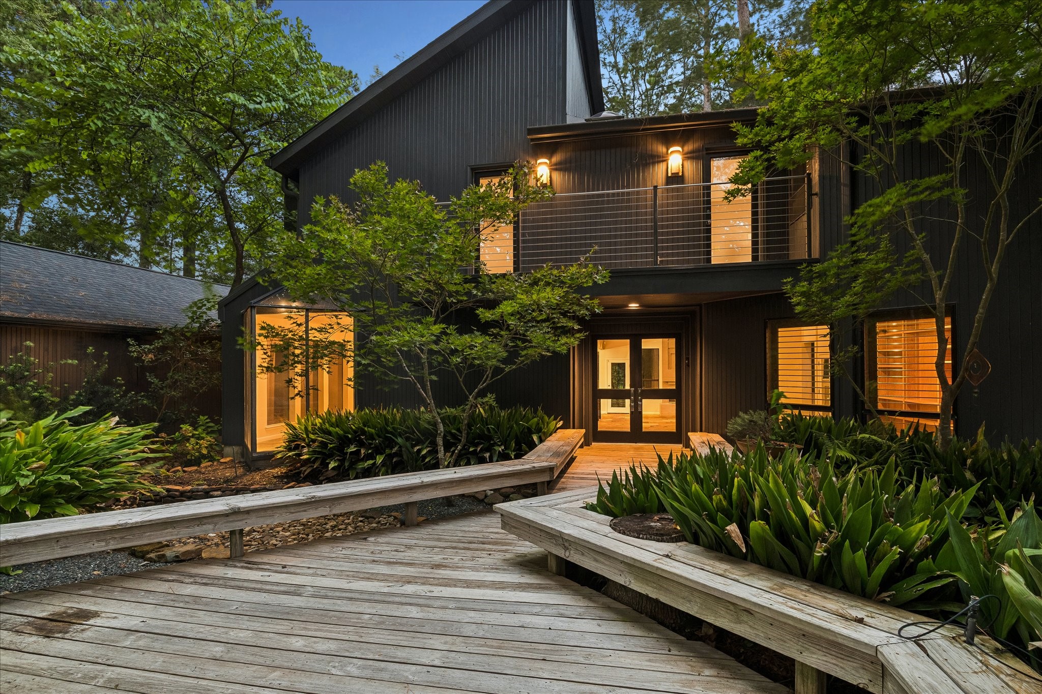 A striking blue-hour arrival: the home's dark vertical siding and soaring gable glow warmly from within, framed by mature trees and a Japanese maple. A wraparound wood boardwalk winds through lush landscaping to the double-door entry, while the cable-railed upper balcony hints at the architectural drama inside. A striking blue-hour arrival: the home's dark vertical siding and soaring gable glow warmly from within, framed by mature trees and a Japanese maple. A wraparound wood boardwalk winds through lush landscaping to the double-door entry, while the cable-railed upper balcony hints at the architectural drama inside.