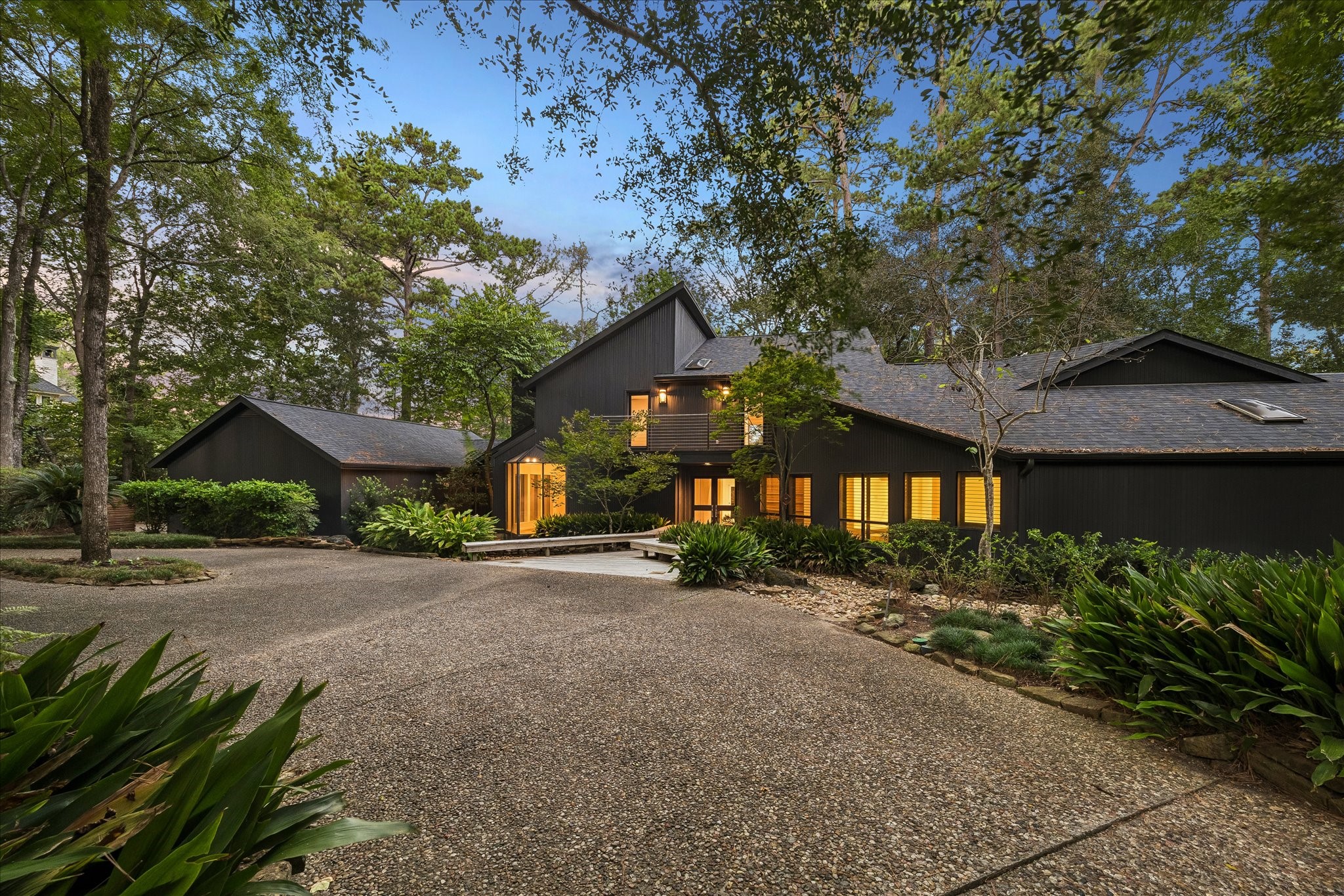 A sweeping gravel drive curves through towering pines and lush understory plantings toward this striking modern home. Multiple gable rooflines and board-formed siding create bold architectural silhouettes against the evening sky, while a wooden boardwalk guides guests to the illuminated entrance total privacy just steps from the front door. A sweeping gravel drive curves through towering pines and lush understory plantings toward this striking modern home. Multiple gable rooflines and board-formed siding create bold architectural silhouettes against the evening sky, while a wooden boardwalk guides guests to the illuminated entrance total privacy just steps from the front door.
