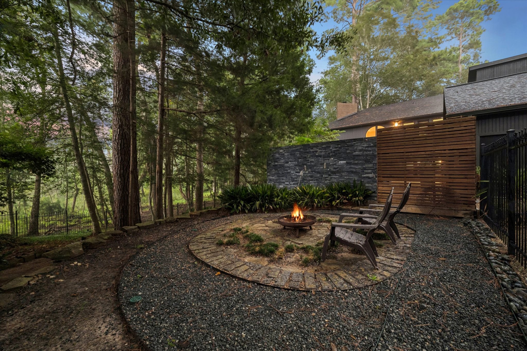 Tucked into the wooded backyard, a circular stone fire pit patio offers an intimate gathering spot surrounded by native plantings and a dramatic slate stone wall. Adirondack chairs invite quiet evenings by the fire, with a sleek wood-slat privacy screen adding modern texture against the natural setting. Tucked into the wooded backyard, a circular stone fire pit patio offers an intimate gathering spot surrounded by native plantings and a dramatic slate stone wall. Adirondack chairs invite quiet evenings by the fire, with a sleek wood-slat privacy screen adding modern texture against the natural setting.