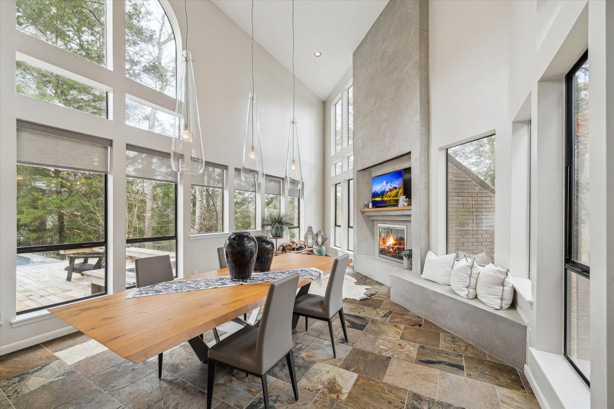Dramatic, light-filled casual dining space featuring soaring ceilings and expansive windows that frame serene wooded views. The open-concept design is highlighted by floor-to-ceiling glass, sleek modern lines, and an abundance of natural light throughout.