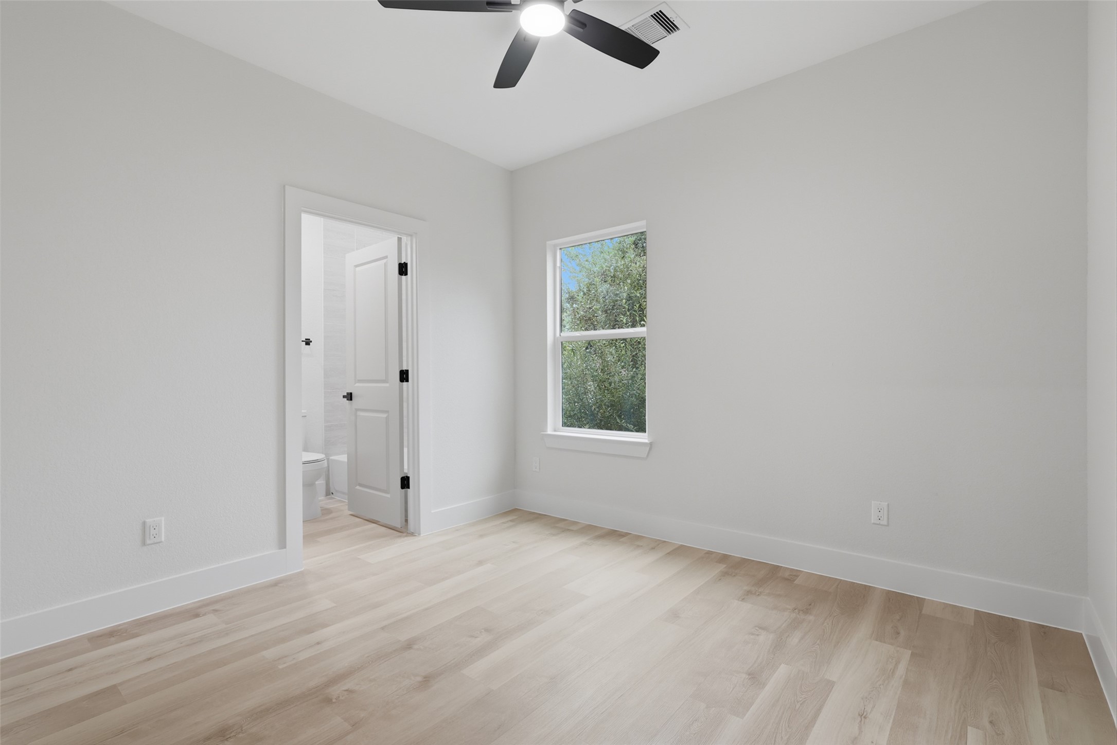 Bright and airy bedroom featuring light wood like LVP flooring, a ceiling fan, and a window with scenic views. Convenient ensuite bathroom access enhances functionality.