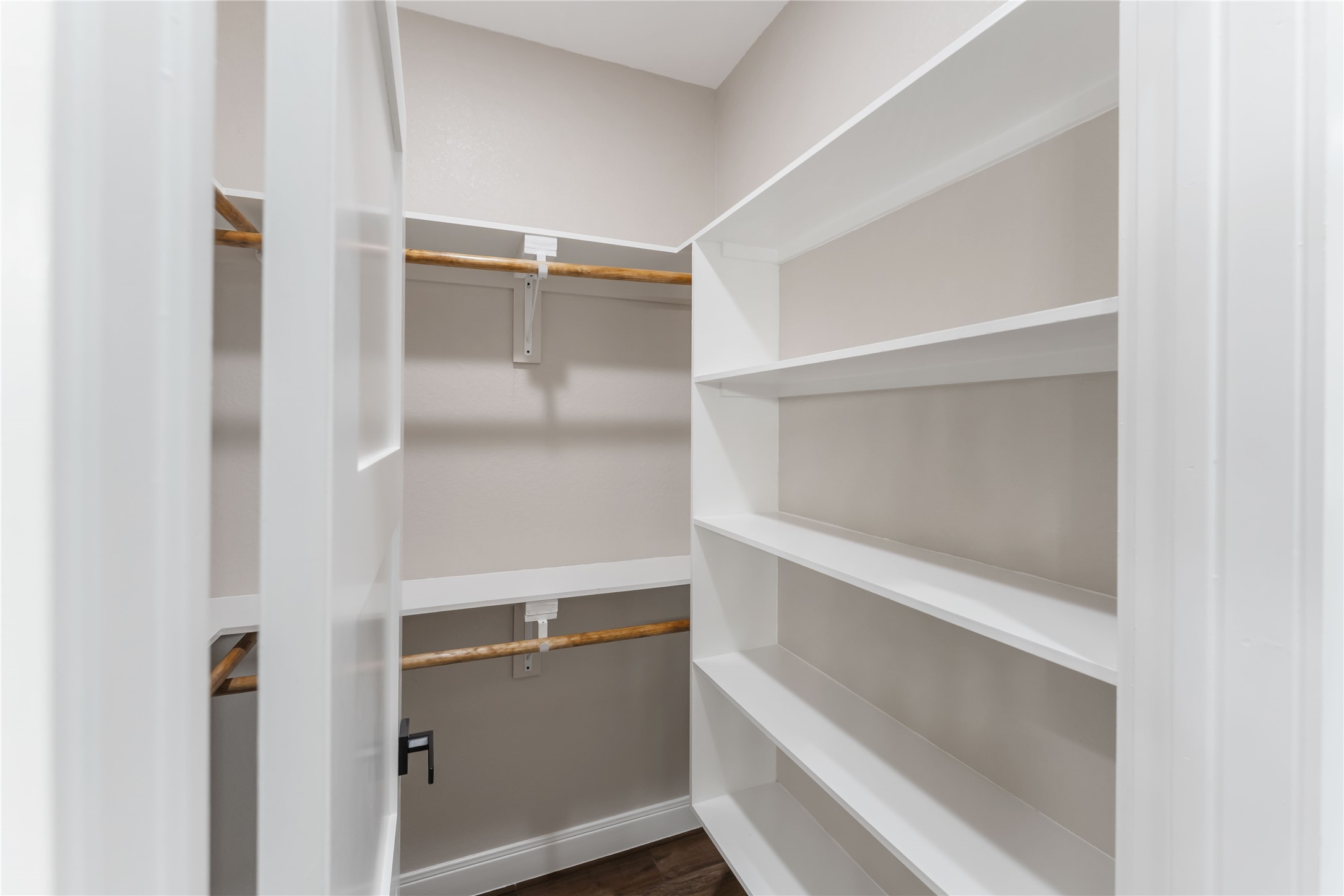 This photo shows a walk-in closet with white shelving on the right and wooden hanging rods on the left. The space is well-organized, providing ample storage for clothes and other items.