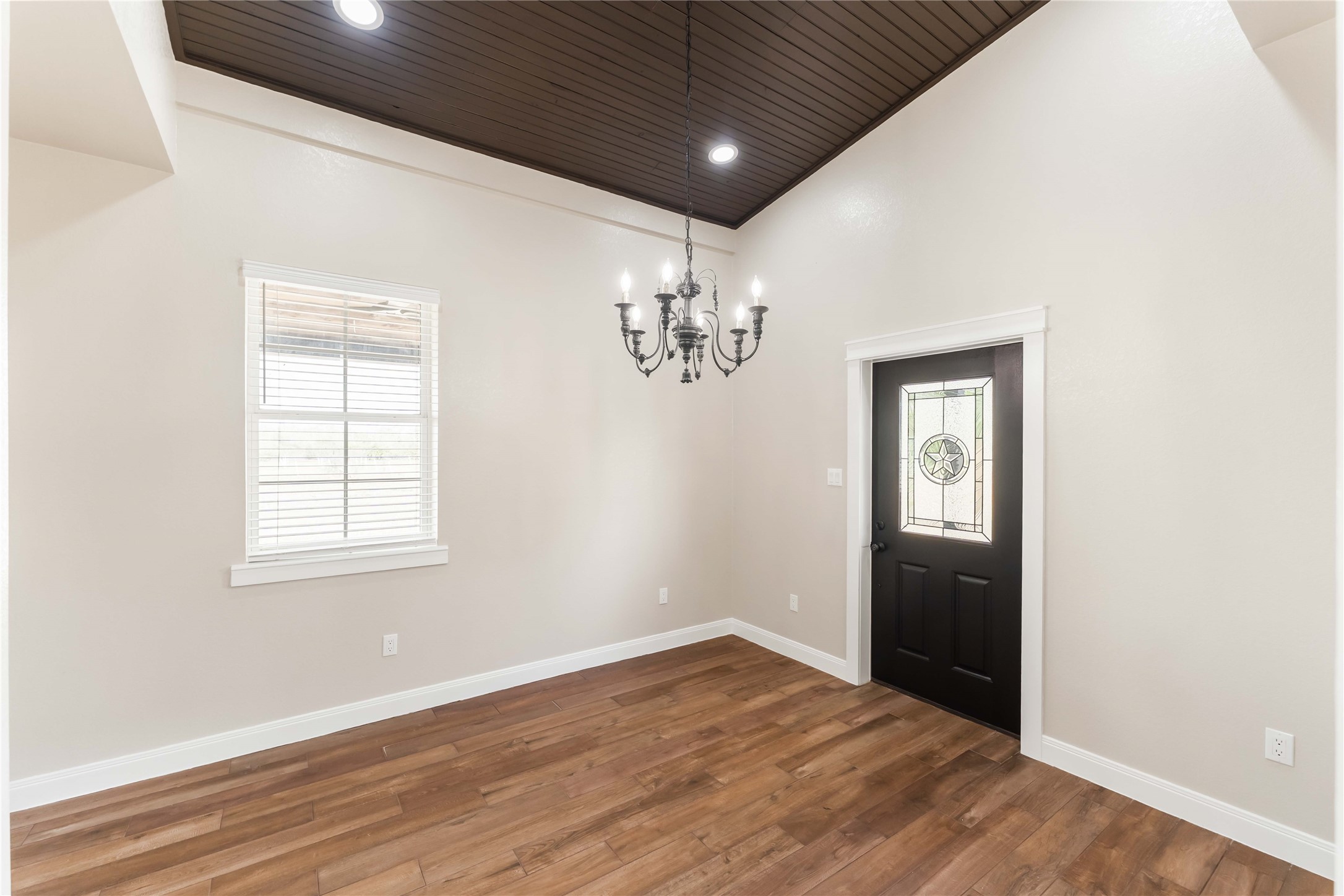 This room features a warm tile wood-style floor and a dark wood ceiling with recessed lighting. It includes a stylish chandelier and a decorative front door with glass detailing, complemented by a window that lets in natural light.