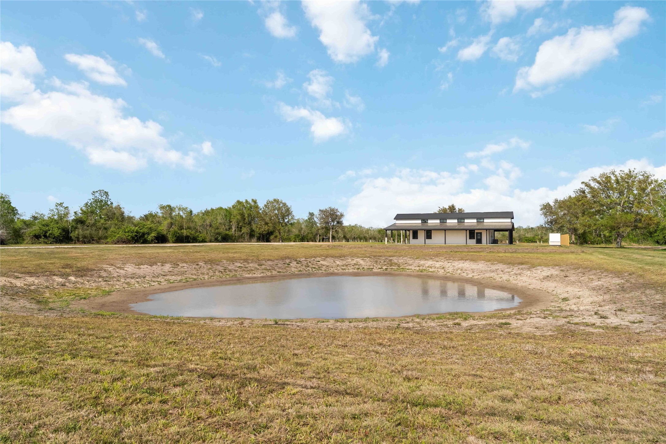 A modern, single-story home is situated on a spacious plot with a small pond in the foreground. The surrounding area is open and grassy, bordered by trees under a clear blue sky.