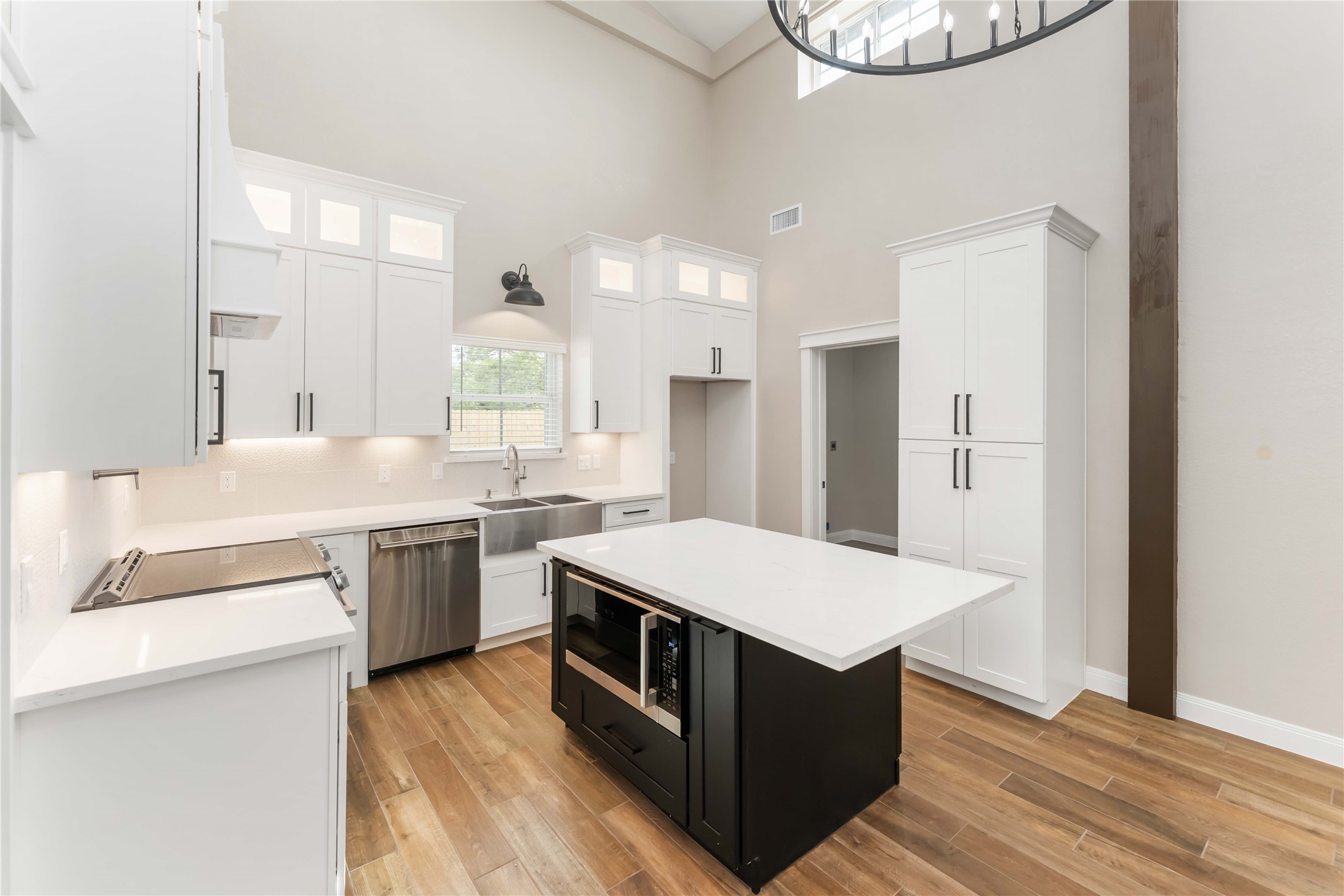 This modern kitchen features high ceilings, white cabinetry, and stainless steel appliances. It includes a central island with a built-in oven and a farmhouse sink. The space is bright with natural light and has tile wood-style flooring.