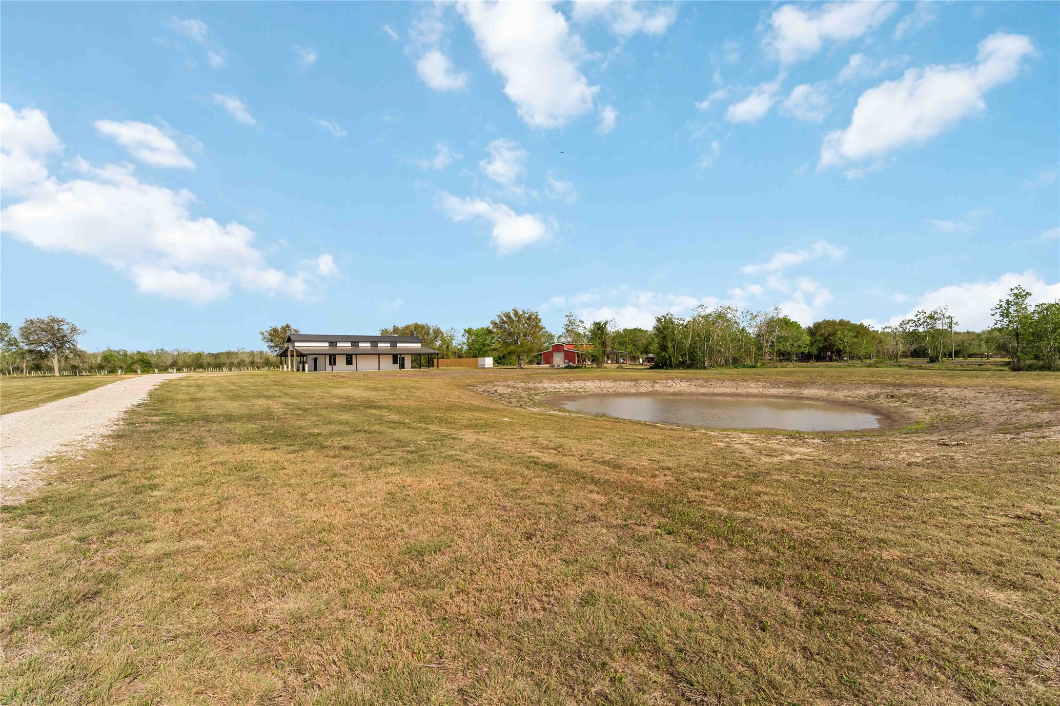 Expansive property with a modern house, a small pond, and a long gravel driveway, surrounded by open land and trees, offering a serene rural setting.