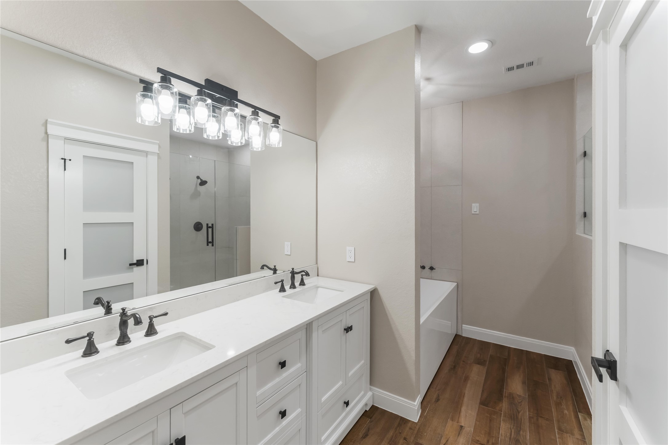 This bathroom features a modern double vanity with sleek black fixtures, a large mirror, and stylish lighting. It includes a spacious glass-enclosed shower and a separate bathtub, all set on elegant tile wood-style flooring.