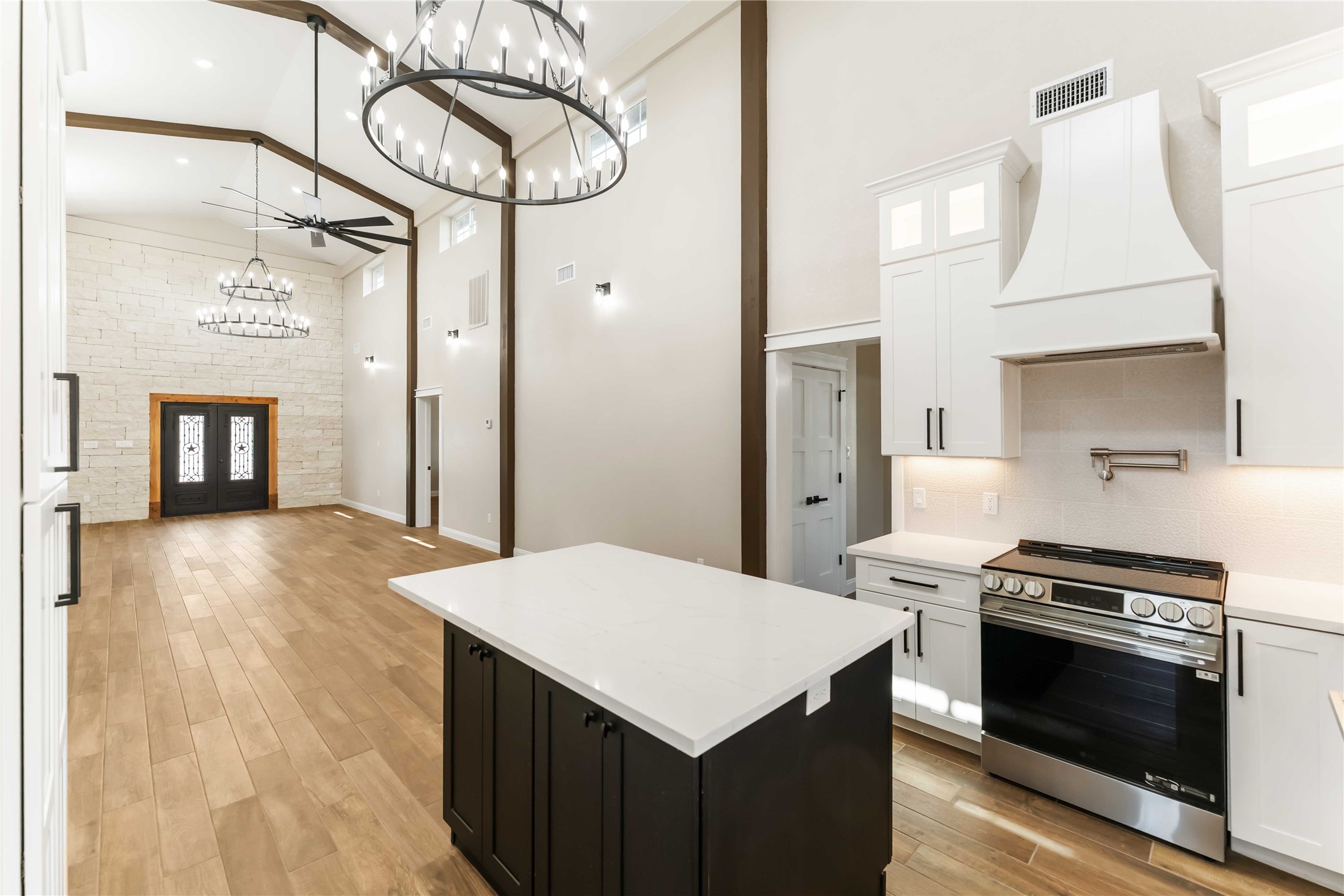 This spacious and elegant open-concept living area features high ceilings, modern chandeliers, and a stone accent wall. The kitchen boasts sleek cabinetry, a central island, and stainless steel appliances, all complemented by warm wood look tile flooring. This spacious and elegant open-concept living area features high ceilings, modern chandeliers, and a stone accent wall. The kitchen boasts sleek cabinetry, a central island, and stainless steel appliances, all complemented by warm wood look tile flooring.