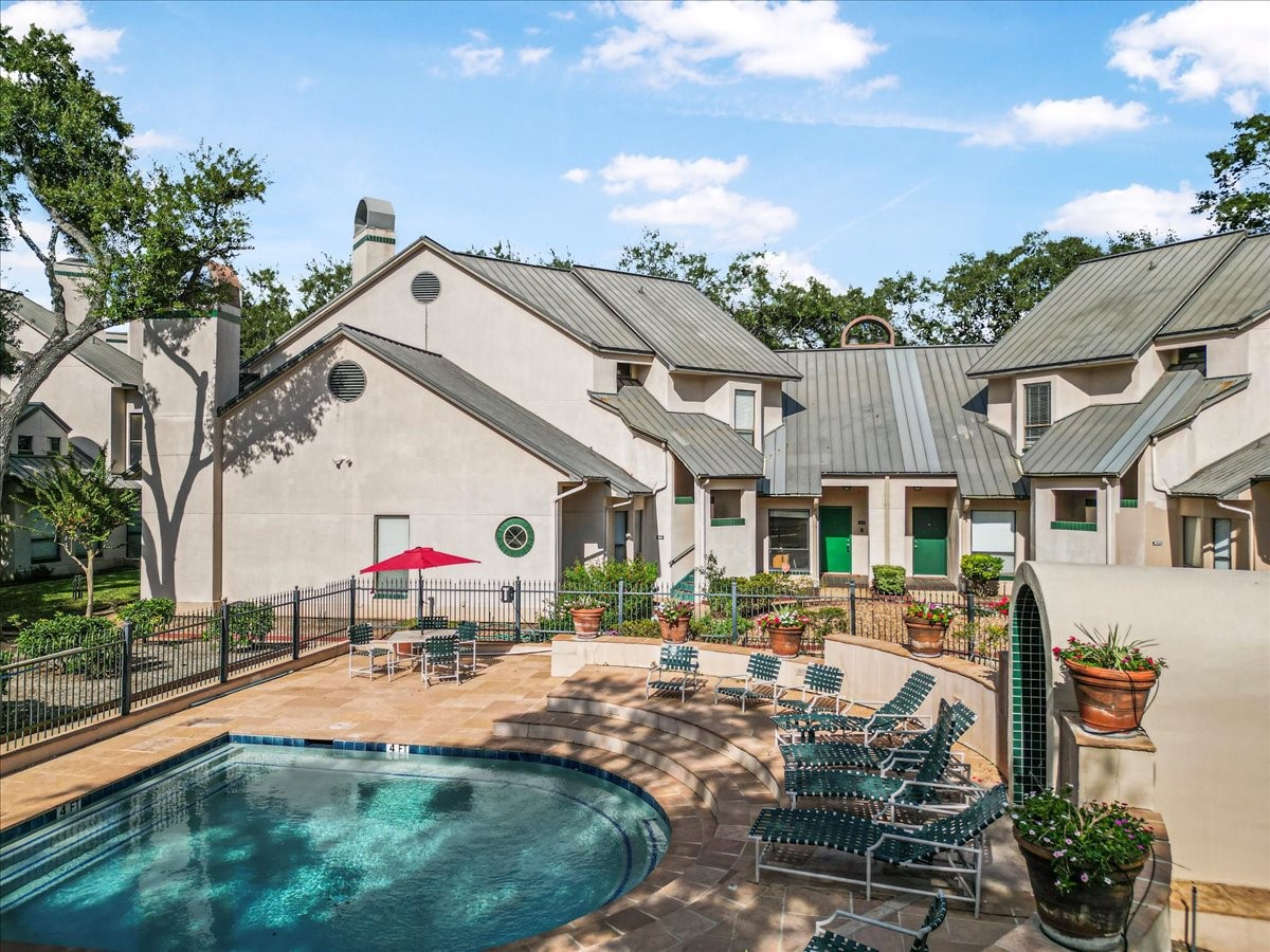 This property features a charming exterior with a metal roof and a shared courtyard. It includes a fenced-in pool area with lounge chairs and a patio table, complemented by lush greenery and potted plants, perfect for relaxation and entertaining.