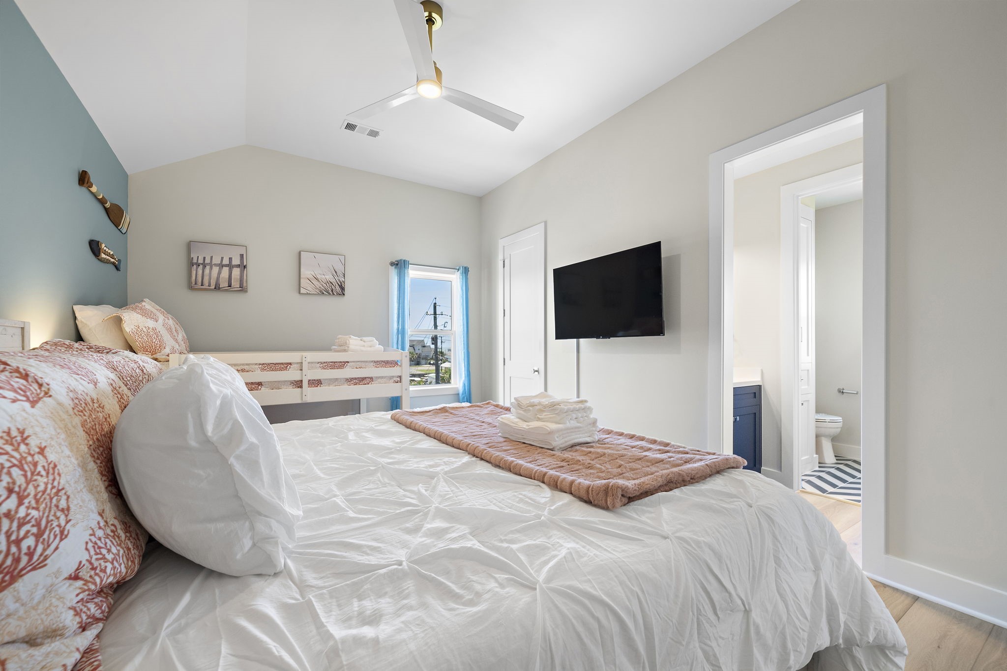 Bright and airy bedroom featuring a comfortable bed with coral-patterned pillows, a wall-mounted TV, and an en-suite bathroom. Perfect for relaxation and comfort.