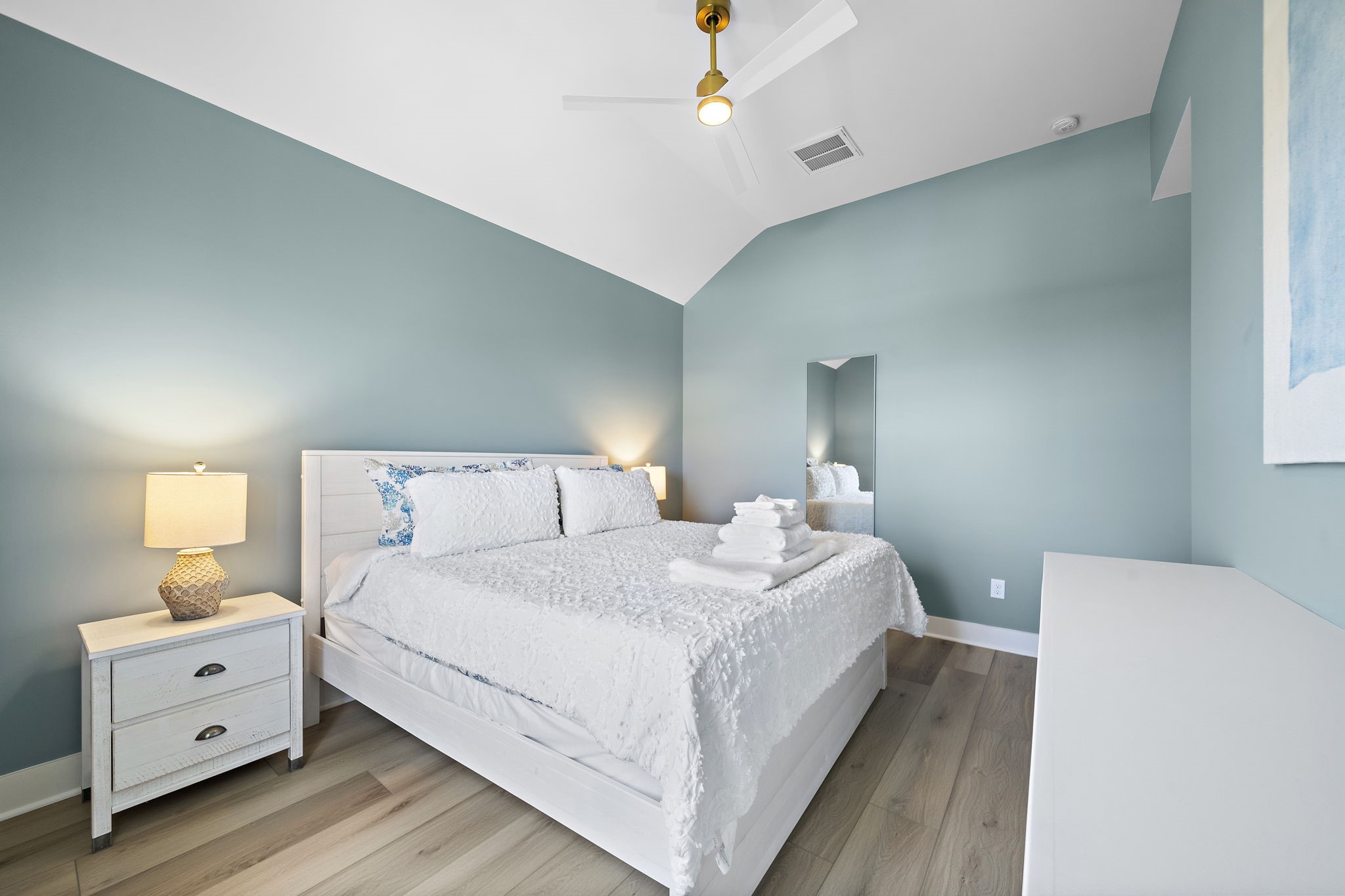 Serene bedroom with soothing blue walls, vaulted ceiling, and elegant wood flooring, featuring a cozy white bed and modern ceiling fan for ultimate comfort.
