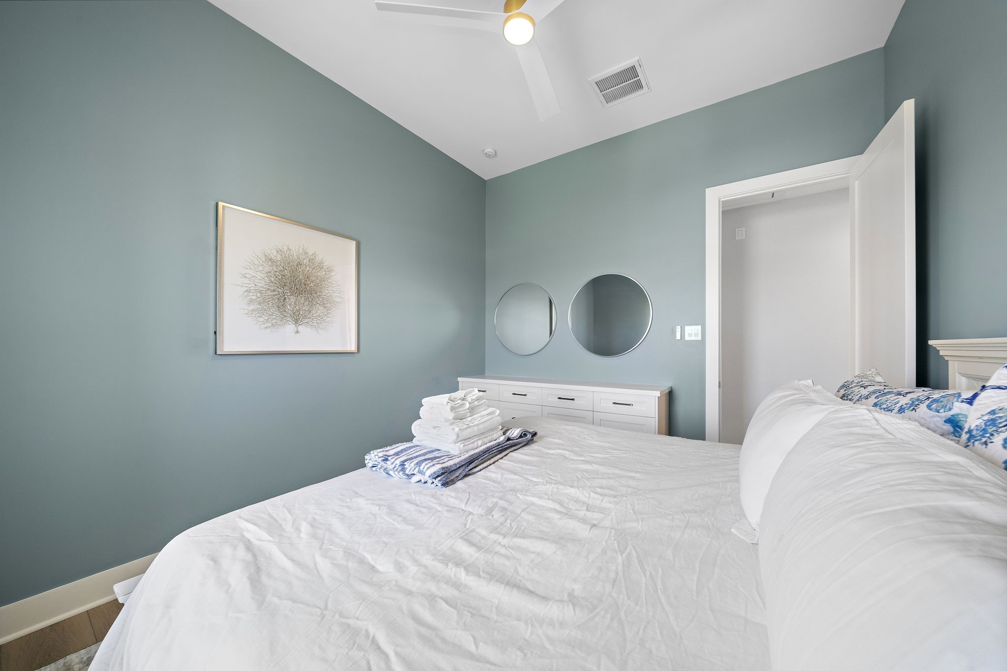 Serene bedroom with soothing blue walls, stylish artwork, and a cozy bed, complemented by a sleek dresser and circular mirrors.