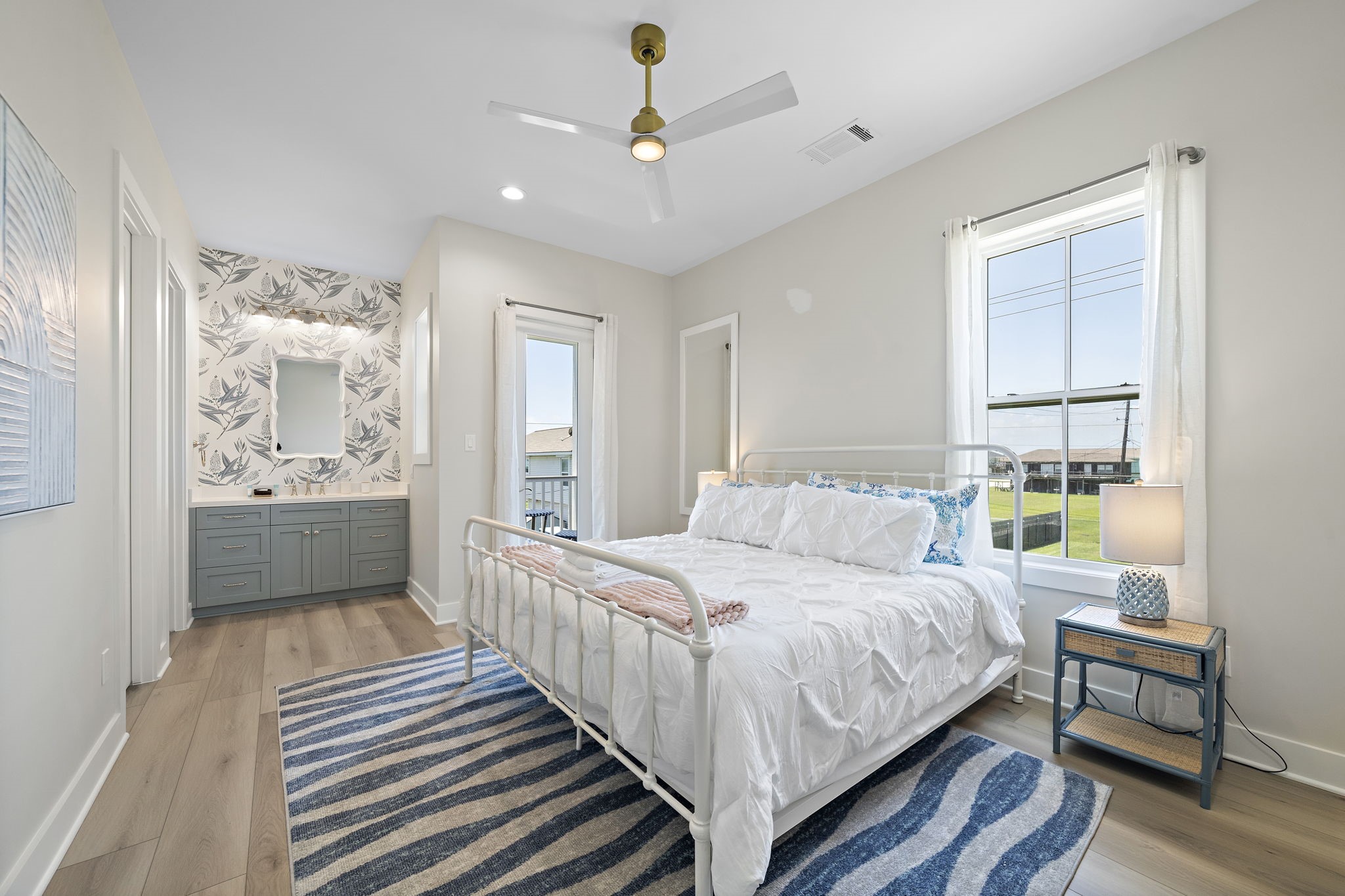 Bright and airy bedroom featuring a cozy bed, stylish ceiling fan, and en-suite bathroom with modern vanity and floral wallpaper. Large windows offer plenty of natural light.