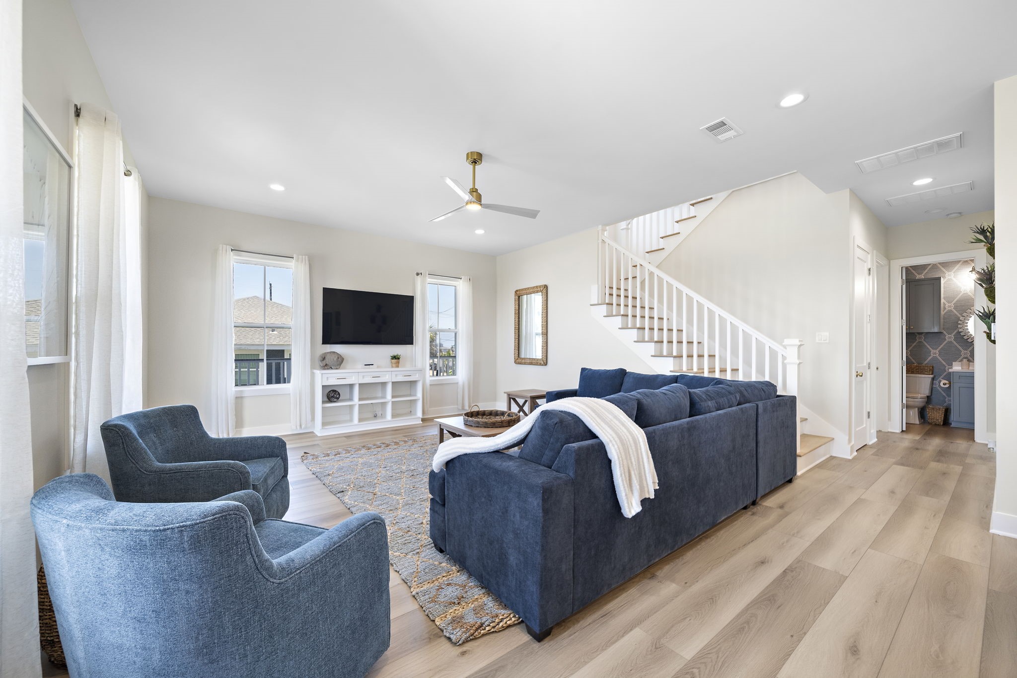Bright and spacious living room featuring elegant hardwood floors, a plush blue sectional, and modern accents, complemented by a stylish ceiling fan and ample natural light. Bright and spacious living room featuring elegant hardwood floors, a plush blue sectional, and modern accents, complemented by a stylish ceiling fan and ample natural light.