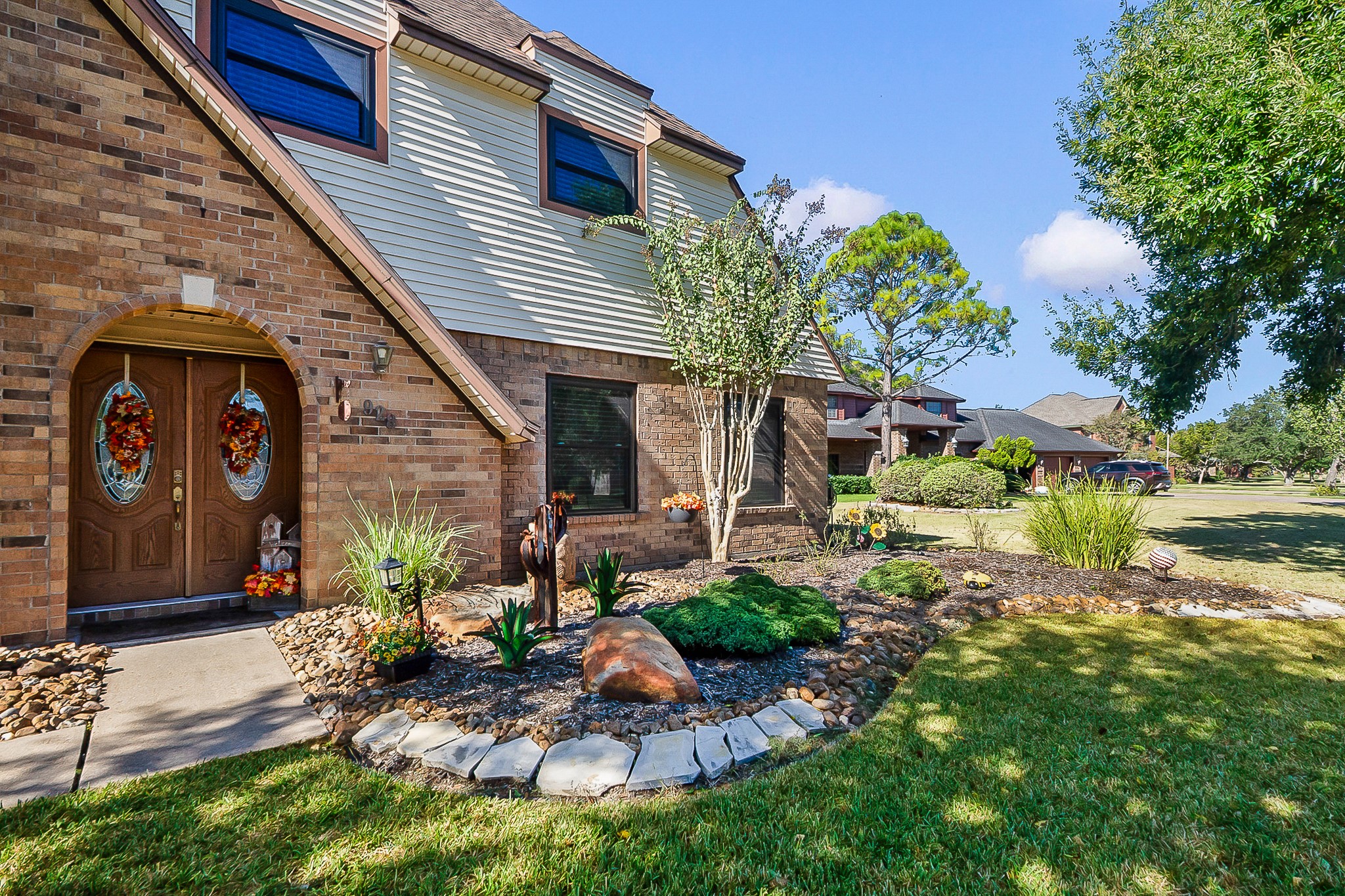 Charming two-story home with a classic brick exterior, welcoming double doors adorned with seasonal decor, and beautifully landscaped front yard featuring lush greenery and stone accents.