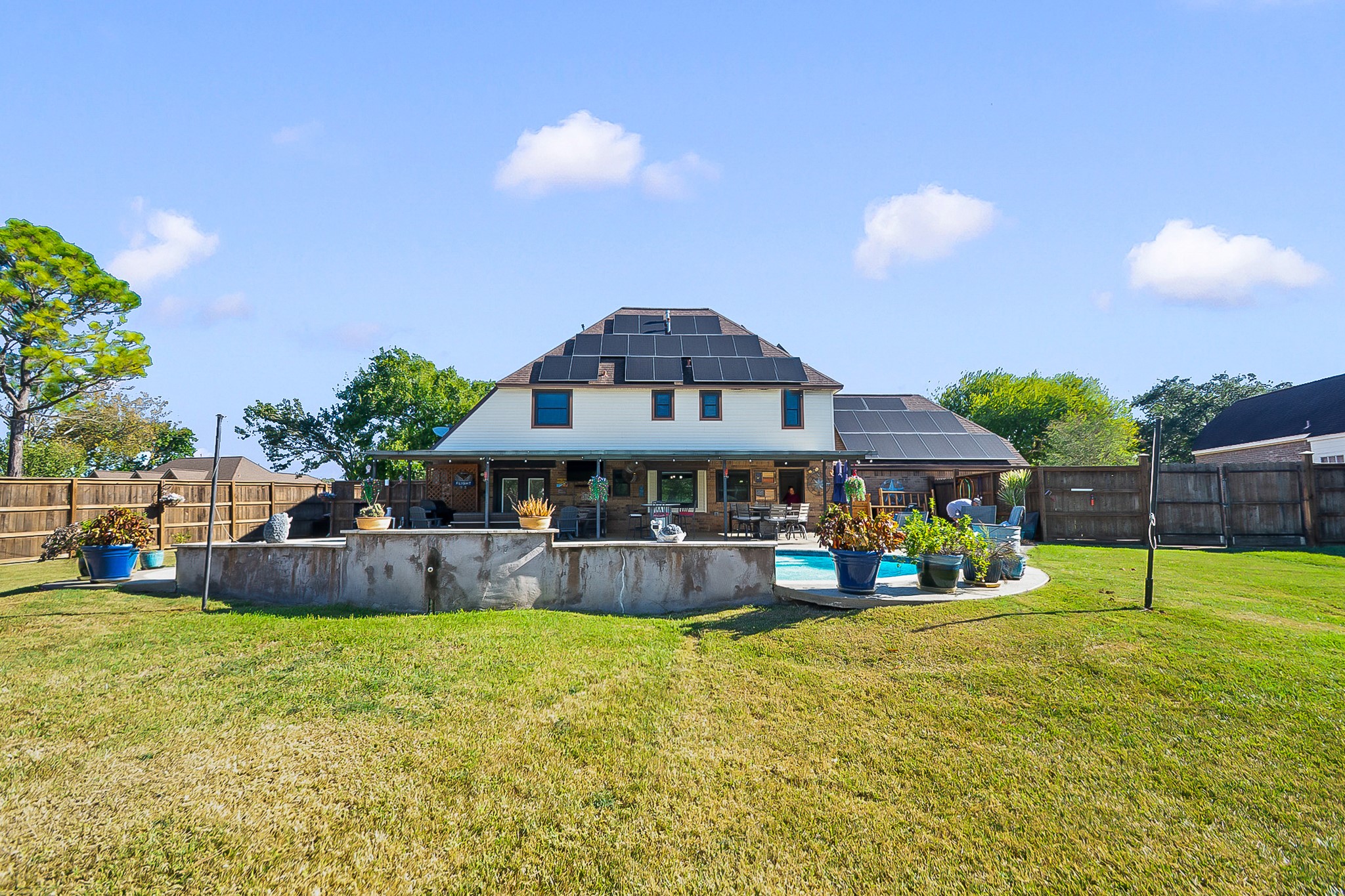 Expansive backyard with lush green lawn, featuring a charming two-story home equipped with paid off solar panels. Enjoy outdoor living with a spacious patio and a well-maintained pool surrounded by potted plants, perfect for relaxation and entertaining.
