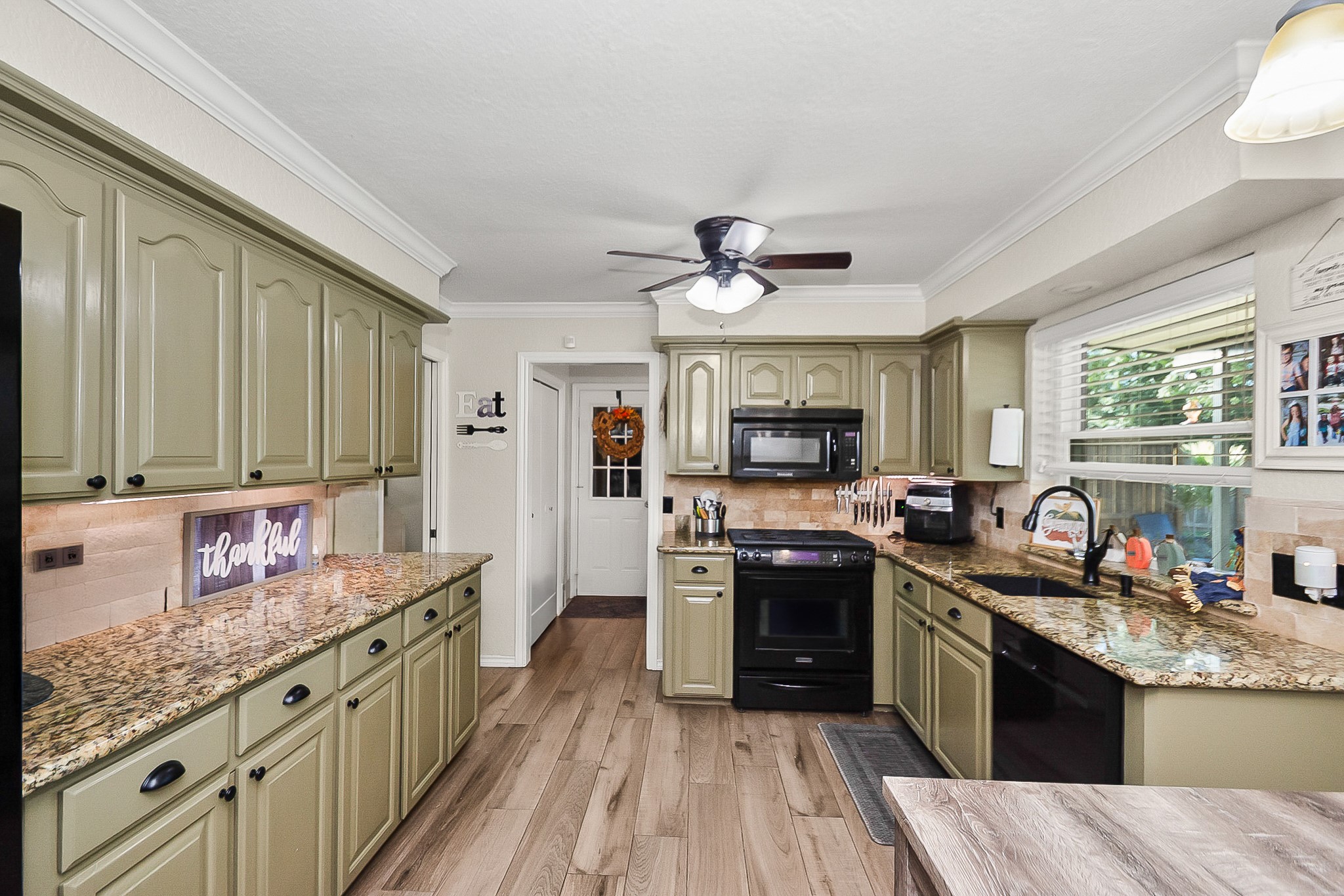 Charming kitchen with olive green cabinetry, sleek granite countertops, and modern black appliances. Natural light streams through the large window, highlighting the spacious layout and warm wood look flooring.