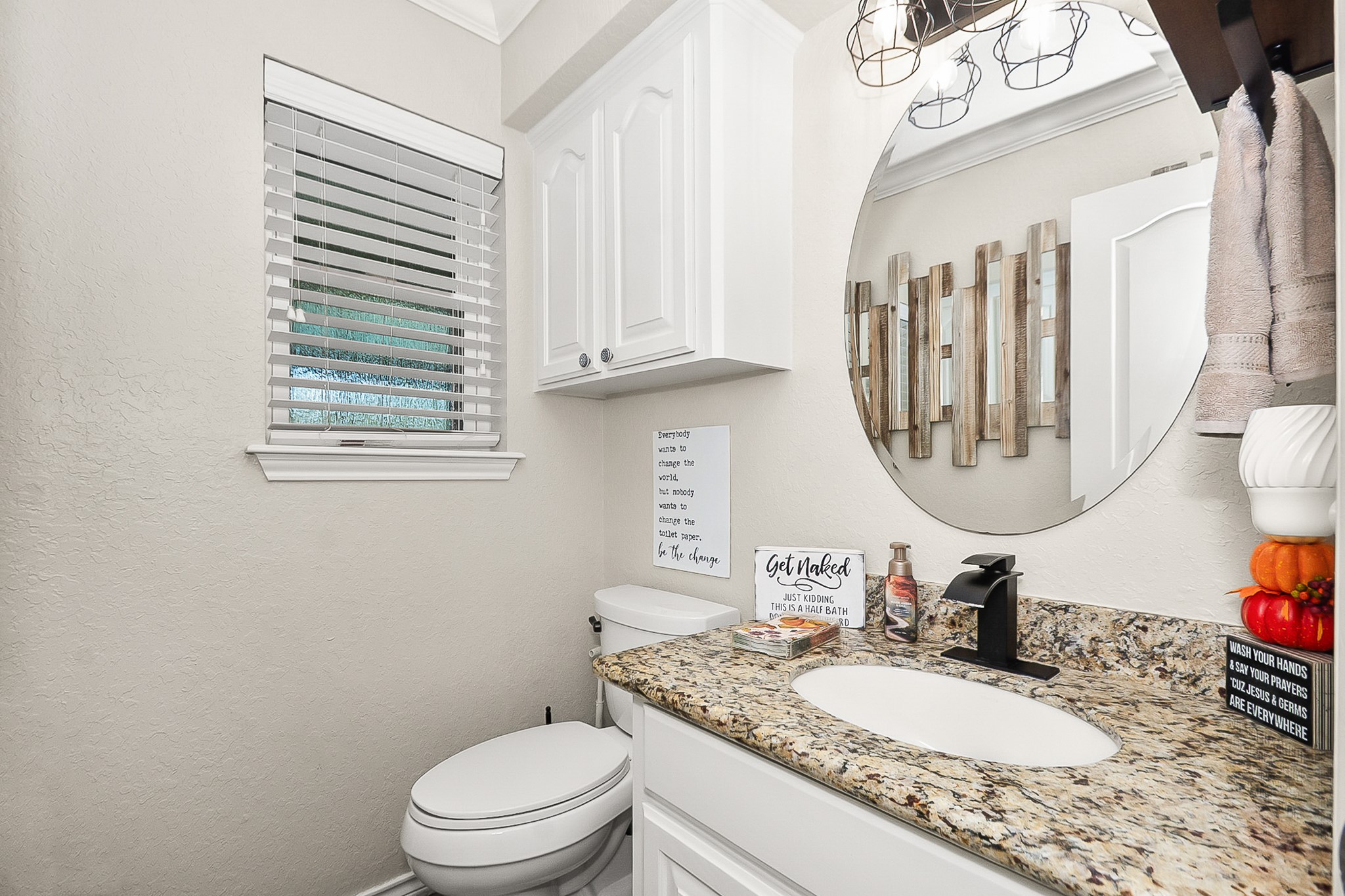 Charming bathroom featuring a granite countertop, round mirror, and sleek black fixtures, enhanced by white cabinetry and a window for natural light.