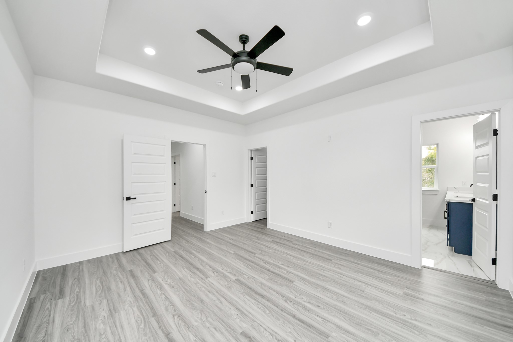 Spacious bedroom featuring elegant tray ceilings, sleek wood flooring, and a modern ceiling fan, with convenient access to an en-suite bathroom.