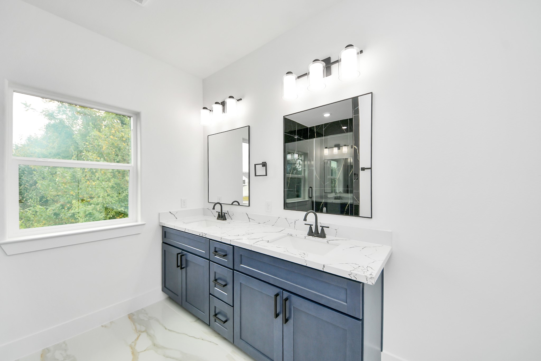 Elegant bathroom featuring a double vanity with sleek blue cabinetry, polished marble countertops, and modern fixtures, complemented by ample natural light from the large window.