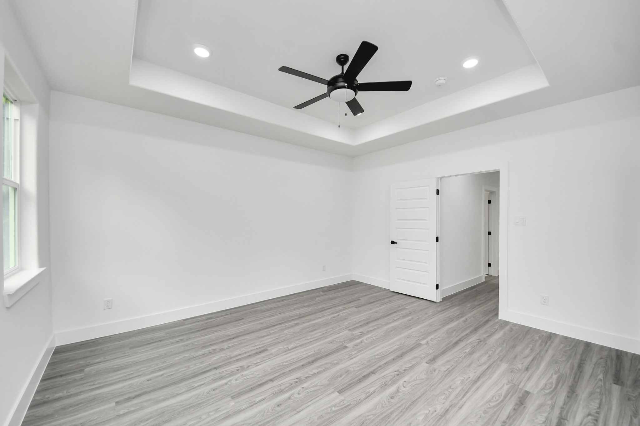 Spacious room featuring elegant tray ceilings, sleek wood flooring, and ample natural light through large windows, complemented by modern recessed lighting and a ceiling fan.