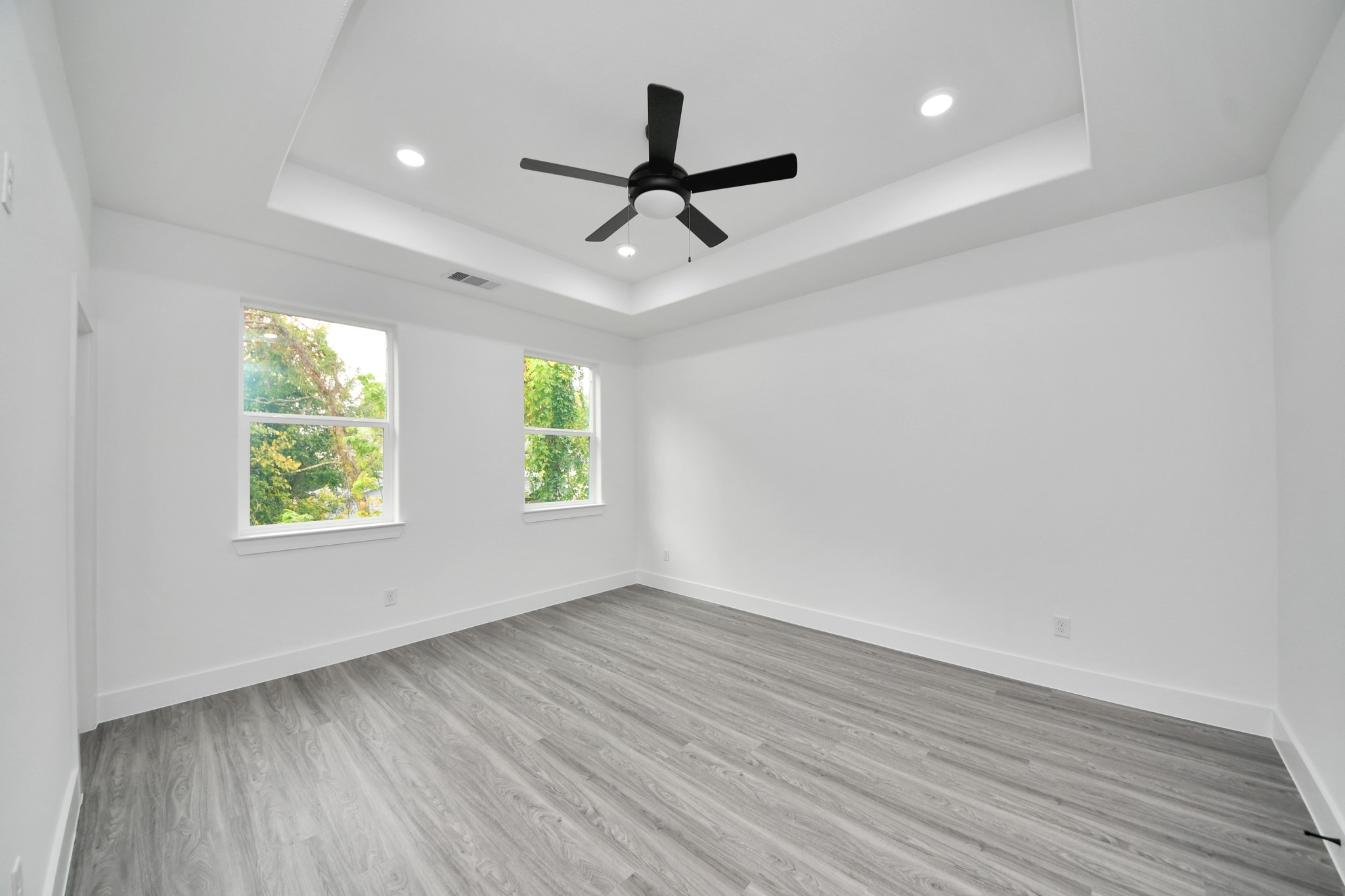 Spacious, light-filled room featuring elegant wood flooring, a stylish tray ceiling with recessed lighting, and a modern ceiling fan, offering a serene and comfortable atmosphere.