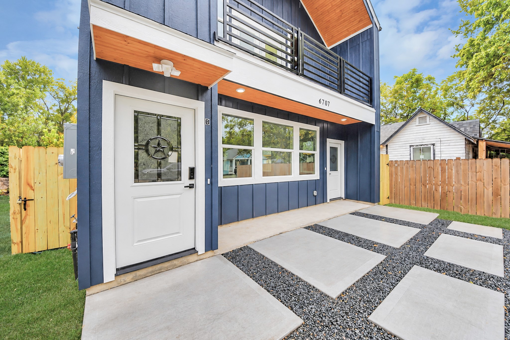 Stylish modern home with a striking navy exterior, accented by white trim and wood paneling. Features include a spacious porch, large windows, and a sleek balcony. The landscaped yard and unique concrete and stone pathway enhance the inviting entrance.