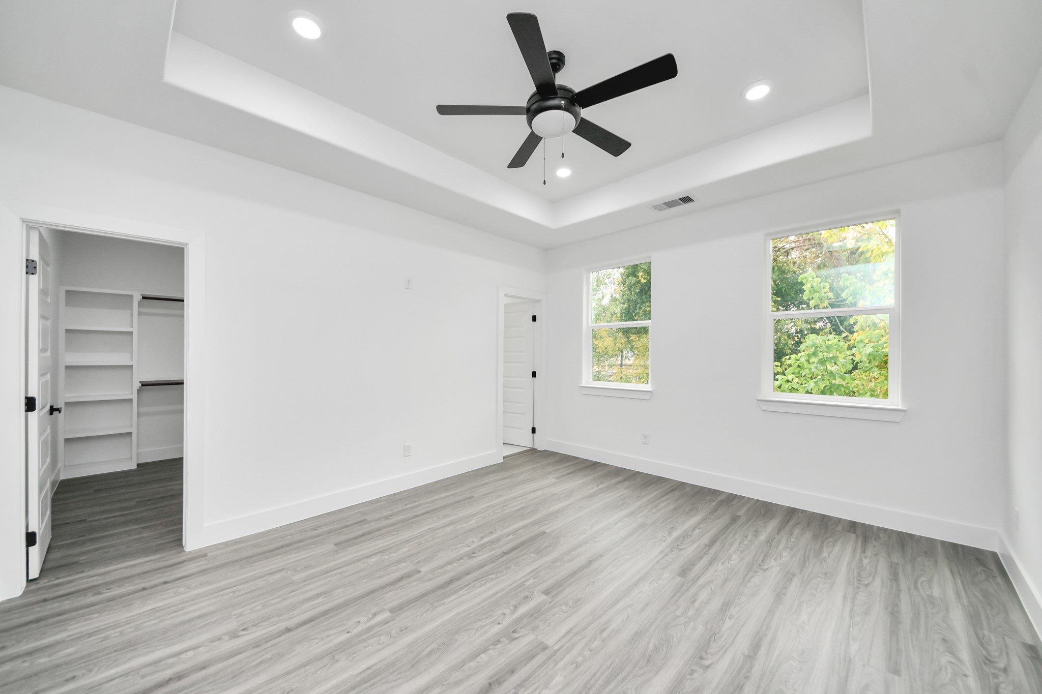 Bright and spacious bedroom featuring a stylish tray ceiling, modern ceiling fan, and large windows that offer abundant natural light. The room includes sleek wood flooring and a generous walk-in closet for ample storage.