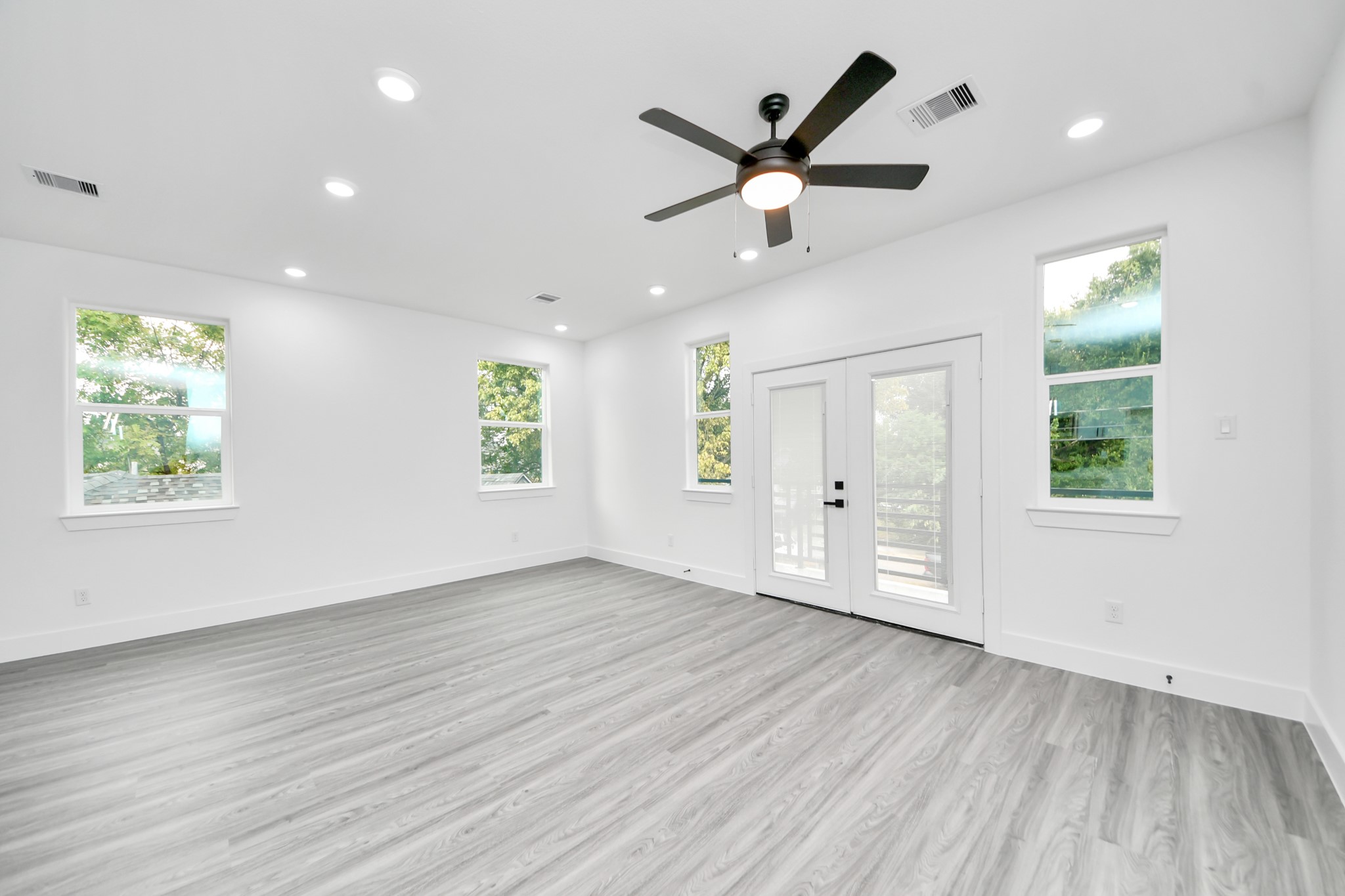 Spacious and bright room featuring sleek wood flooring, French doors leading to an outdoor space, and modern recessed lighting, complemented by a contemporary ceiling fan. Spacious and bright room featuring sleek wood flooring, French doors leading to an outdoor space, and modern recessed lighting, complemented by a contemporary ceiling fan.