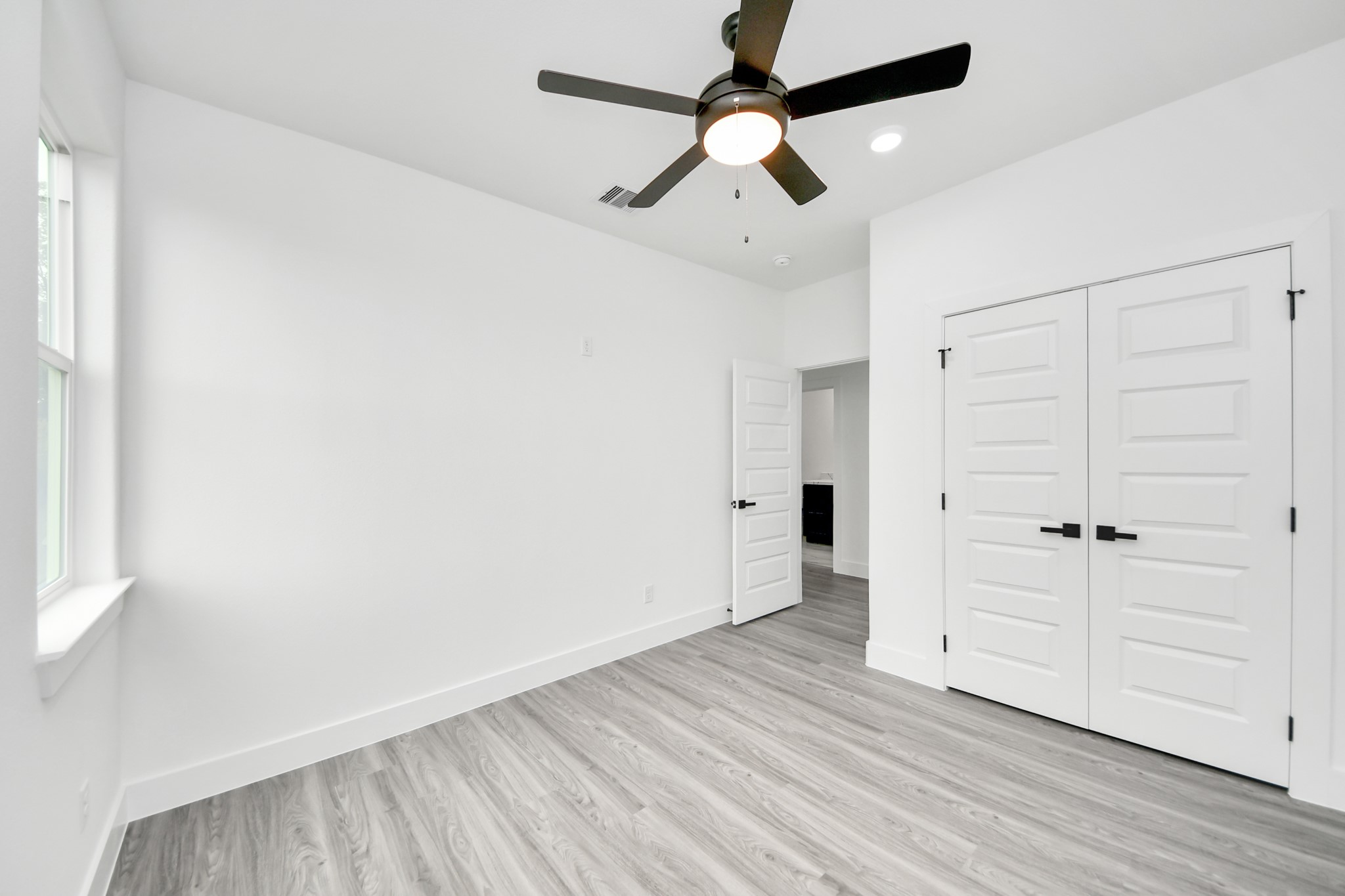 Bright and spacious room featuring sleek wood flooring, a modern ceiling fan, and ample natural light, complemented by crisp white walls and stylish double doors.