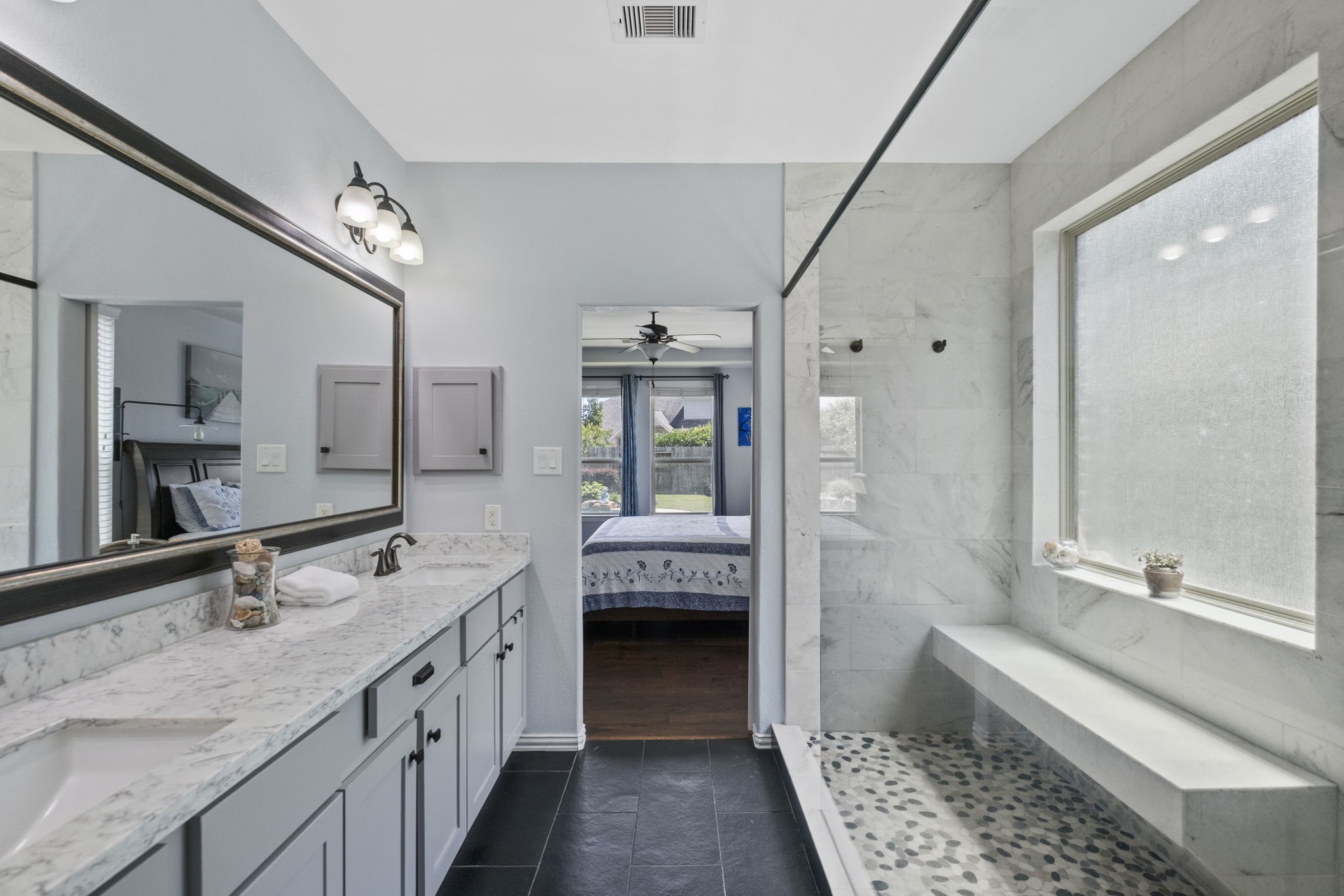 This modern bathroom features a spacious shower with a pebble floor and a built-in bench, a large vanity with dual sinks and quartz countertops, and ample natural light, creating a bright and peaceful space.