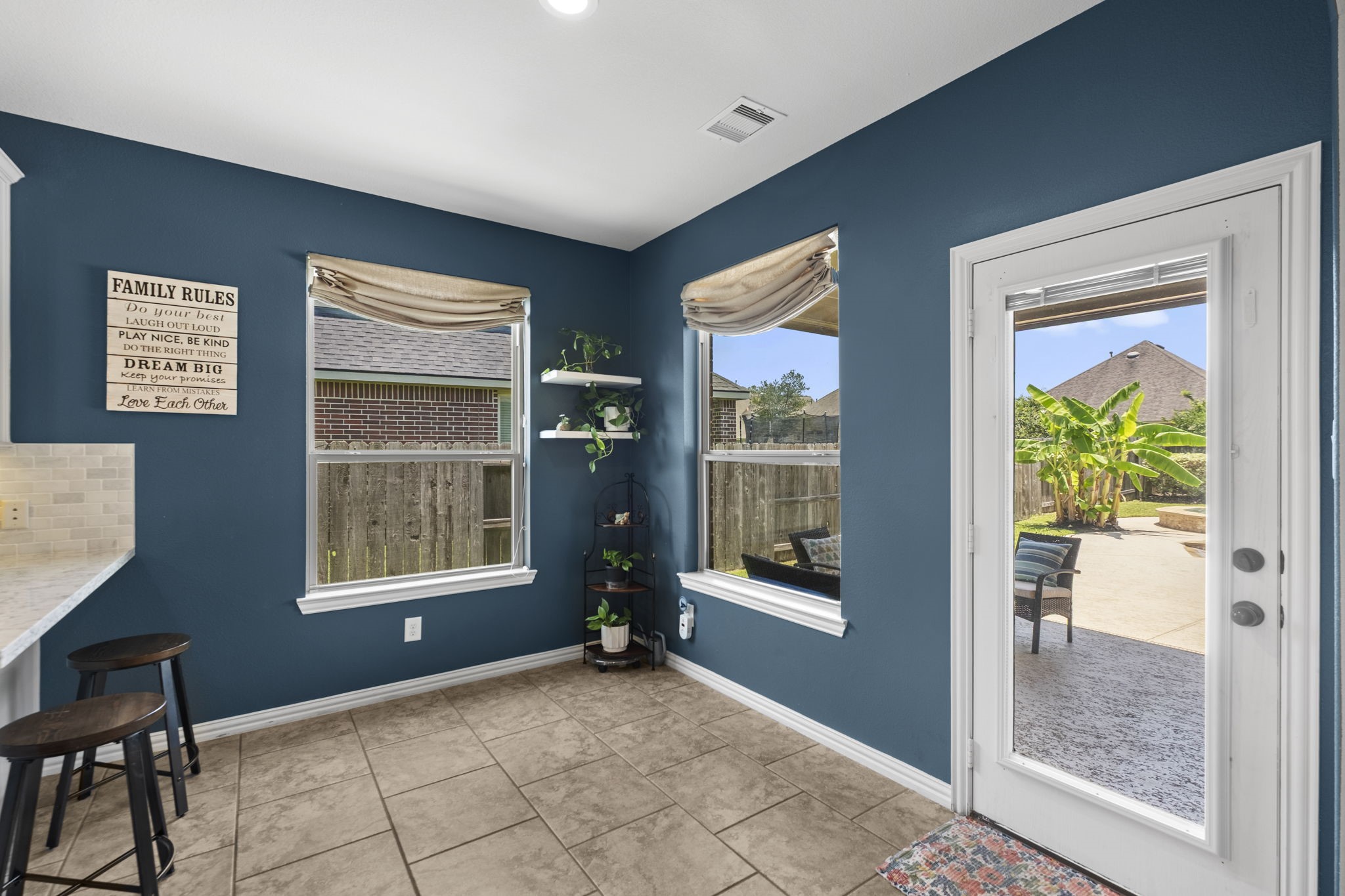 Bright breakfast area with tile flooring, featuring large windows with beige roman shades. A glass door opens to a spacious patio, perfect for outdoor entertaining. A breakfast bar with stools adds convenience, while the decor provides a cozy and inviting atmosphere.