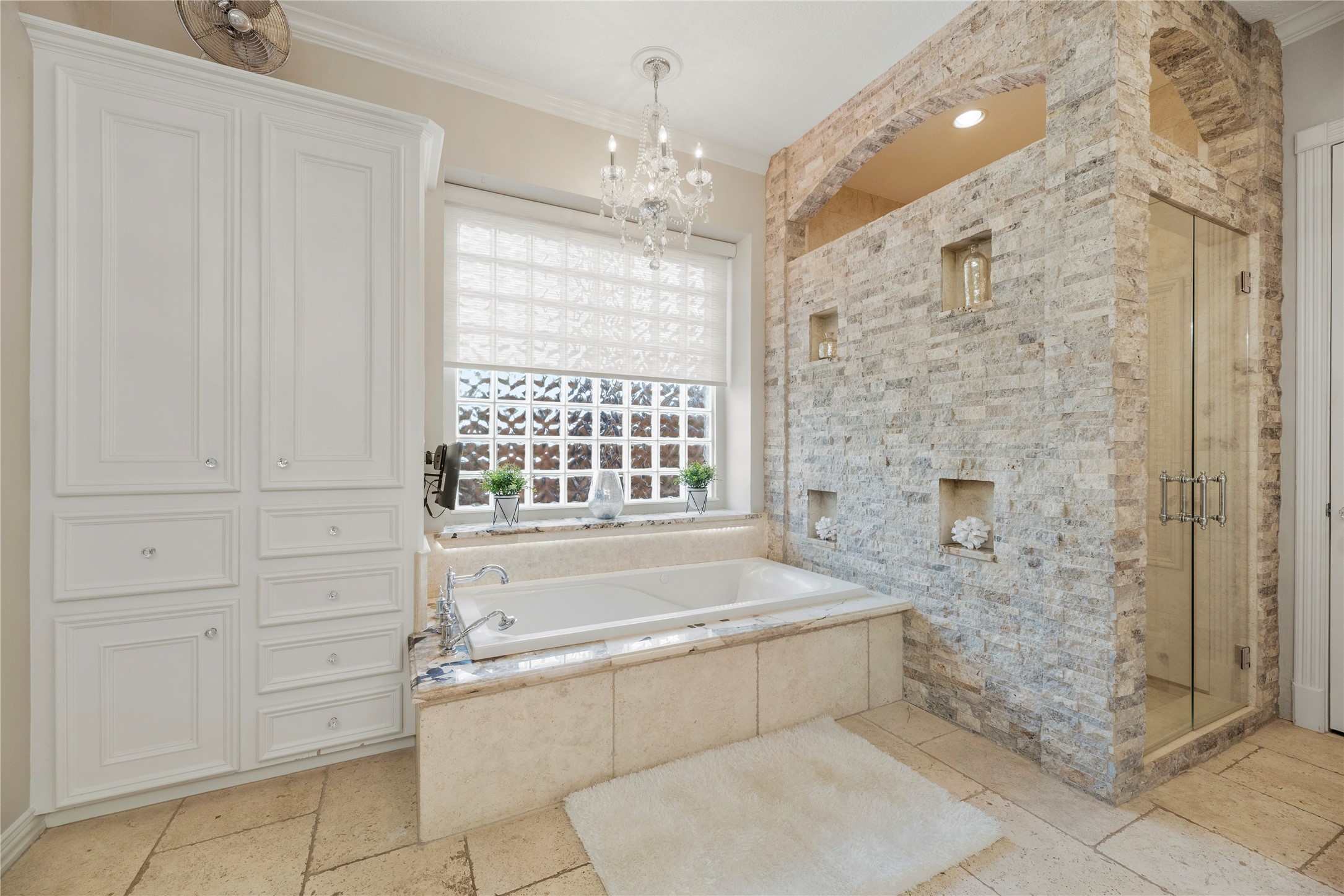 Luxurious primary bathroom features a stunning stone-clad walk-in shower with architectural arches and decorative niches, a generous jetted tub beneath a glass block window, elegant chandelier and extensive custom cabinetry.