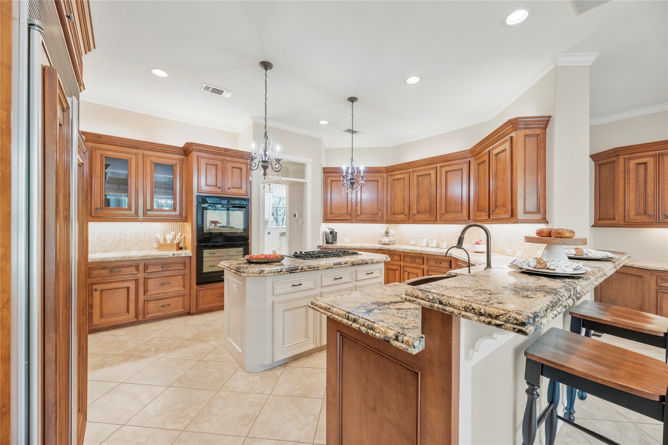 Exquisite gourmet kitchen showcases rich custom-style wood cabinetry, built-in side-by-side refrigerator, granite countertops, elegant tile backsplash, under cabinet lighting, pendant lighting, double ovens and a gas cooktop.