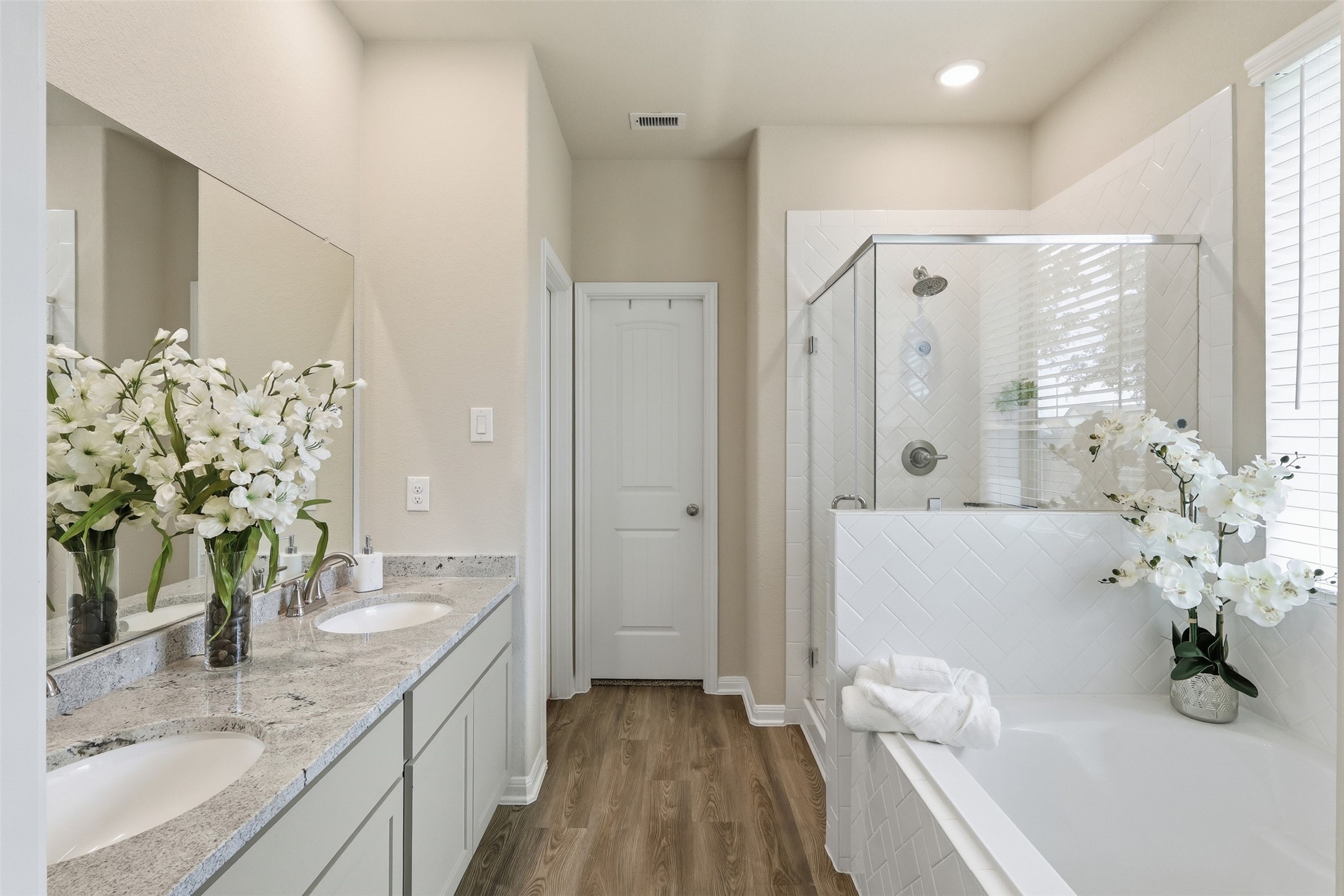 Spa-like primary bath featuring dual granite vanities, a soaking tub, oversized glass-enclosed shower, abundant natural light, and a spacious walk-in closet.