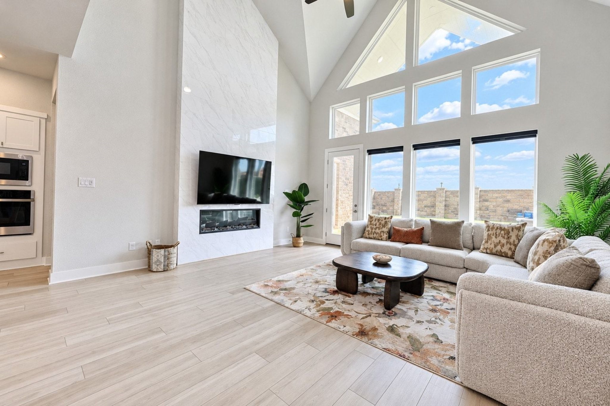 The spacious two-story living room features a soaring vaulted ceiling, a ceiling fan, luxury vinyl plank flooring, and a modern electric fireplace. Expansive windows fill the space with natural light, creating a bright and inviting atmosphere.
