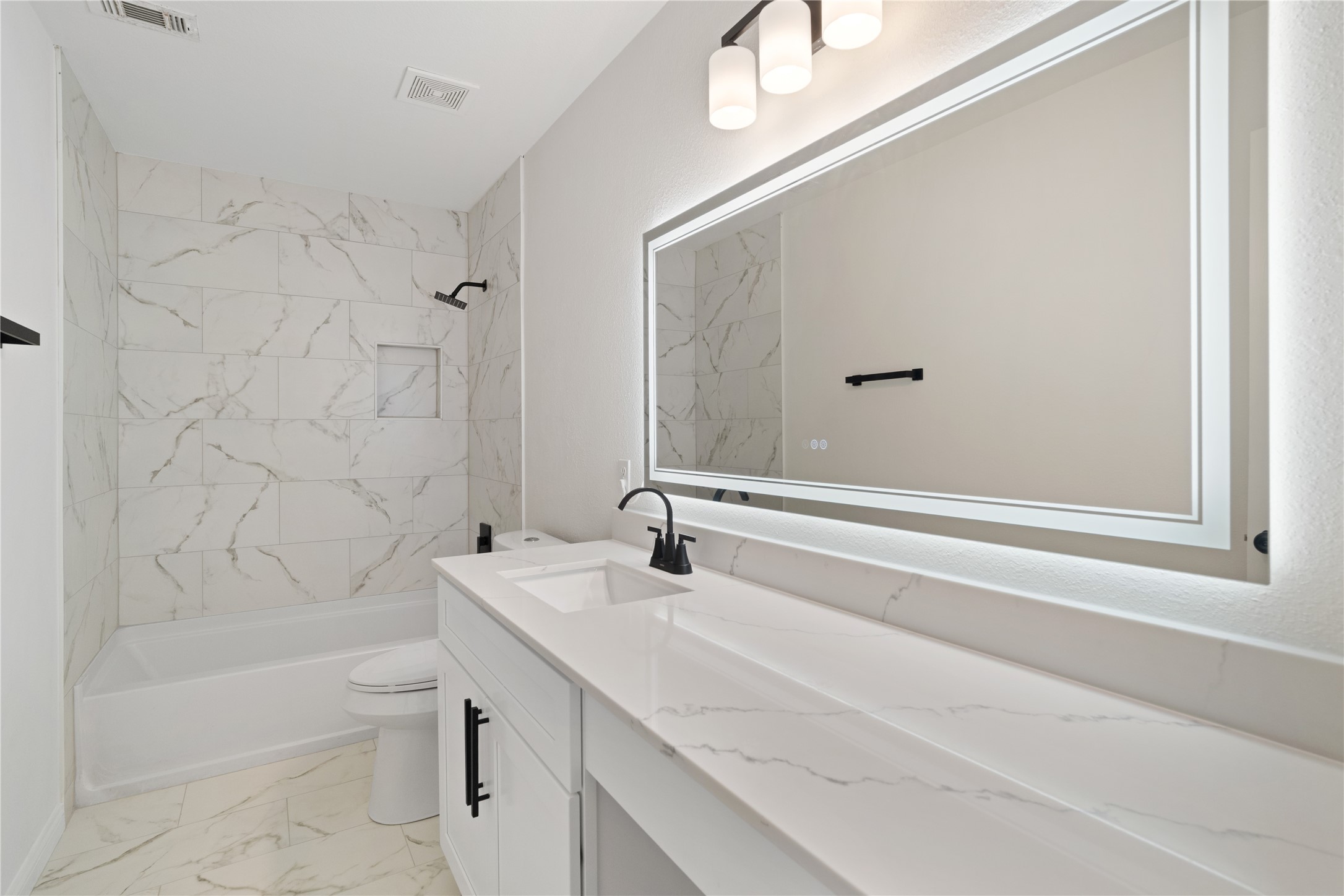 Full Bathroom  Updated bathroom featuring a long vanity with stone-look countertop, illuminated mirror, matte-black fixtures, and a tub/shower surrounded by marble-look tile.