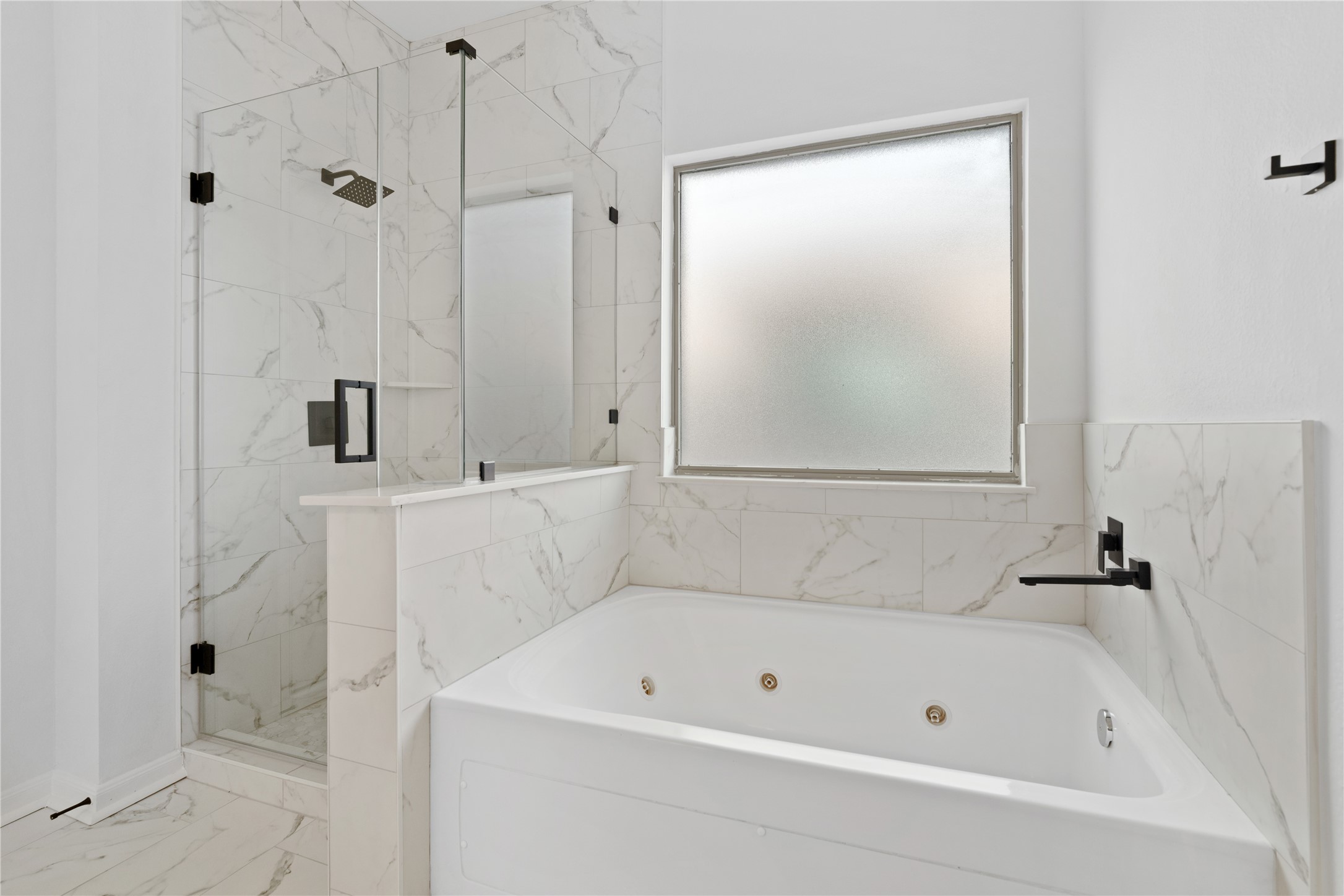 Primary Bathroom  Relaxing bath area features a jetted soaking tub, separate glass-enclosed shower, marble-look tile surrounds, matte-black fixtures, and a privacy-glass window.