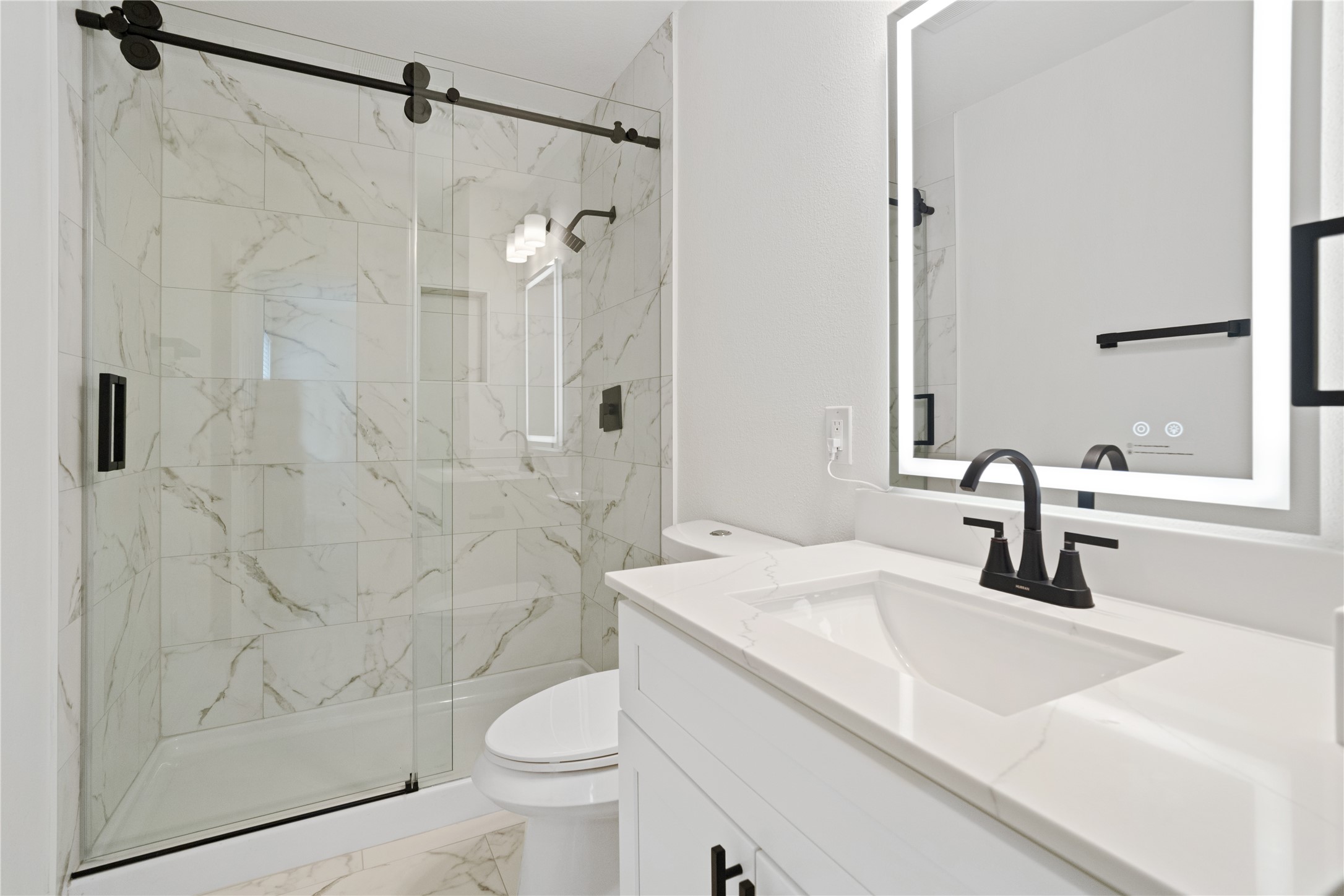 Full Bathroom  Stylish bathroom with marble-look shower tile, a glass sliding enclosure, stone-look vanity top, illuminated mirror, and contrasting matte-black fixtures.
