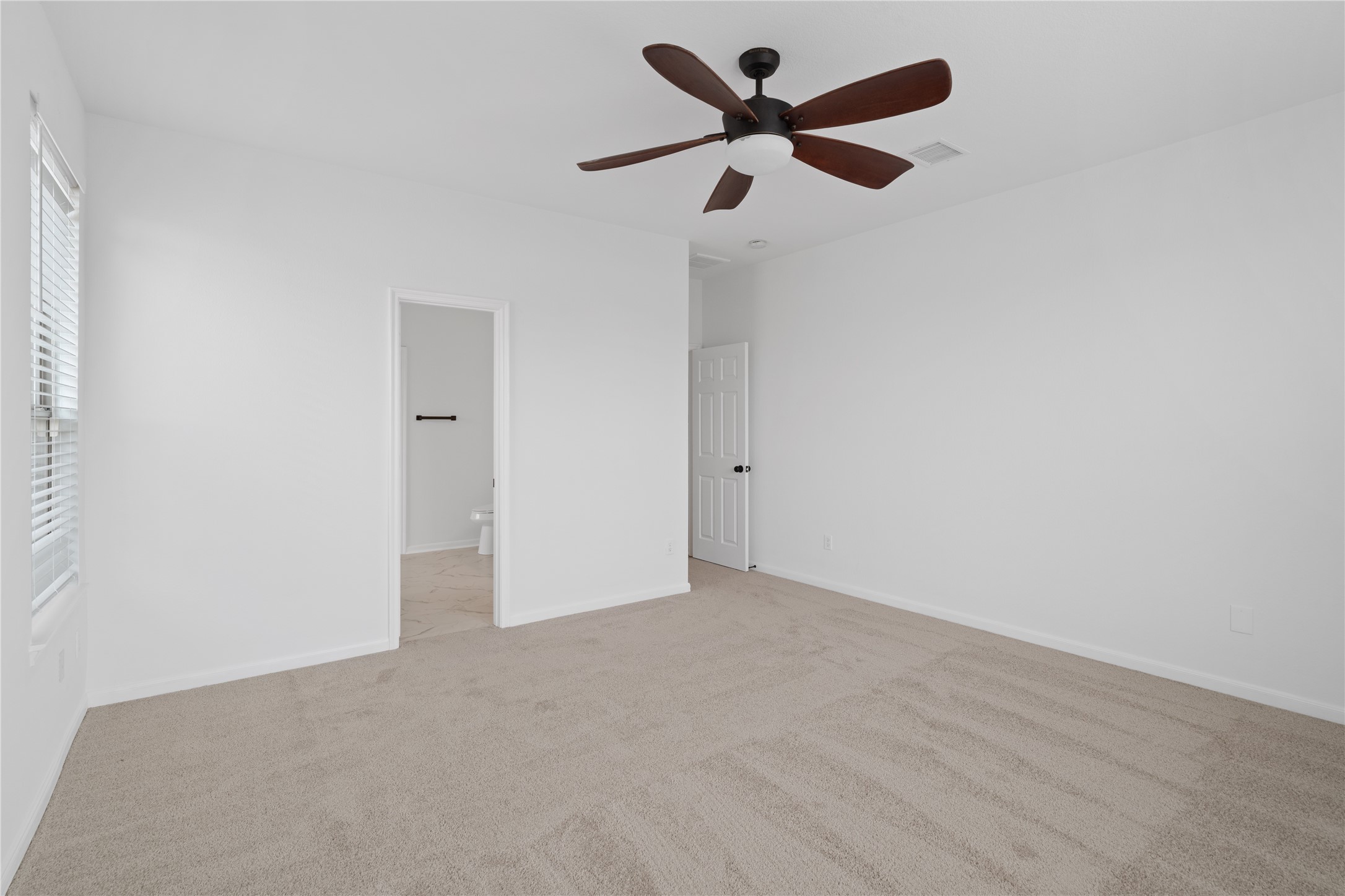 Primary Bedroom  Generously proportioned primary bedroom featuring soft neutral carpet, a ceiling fan, and convenient access to the adjoining bathroom.