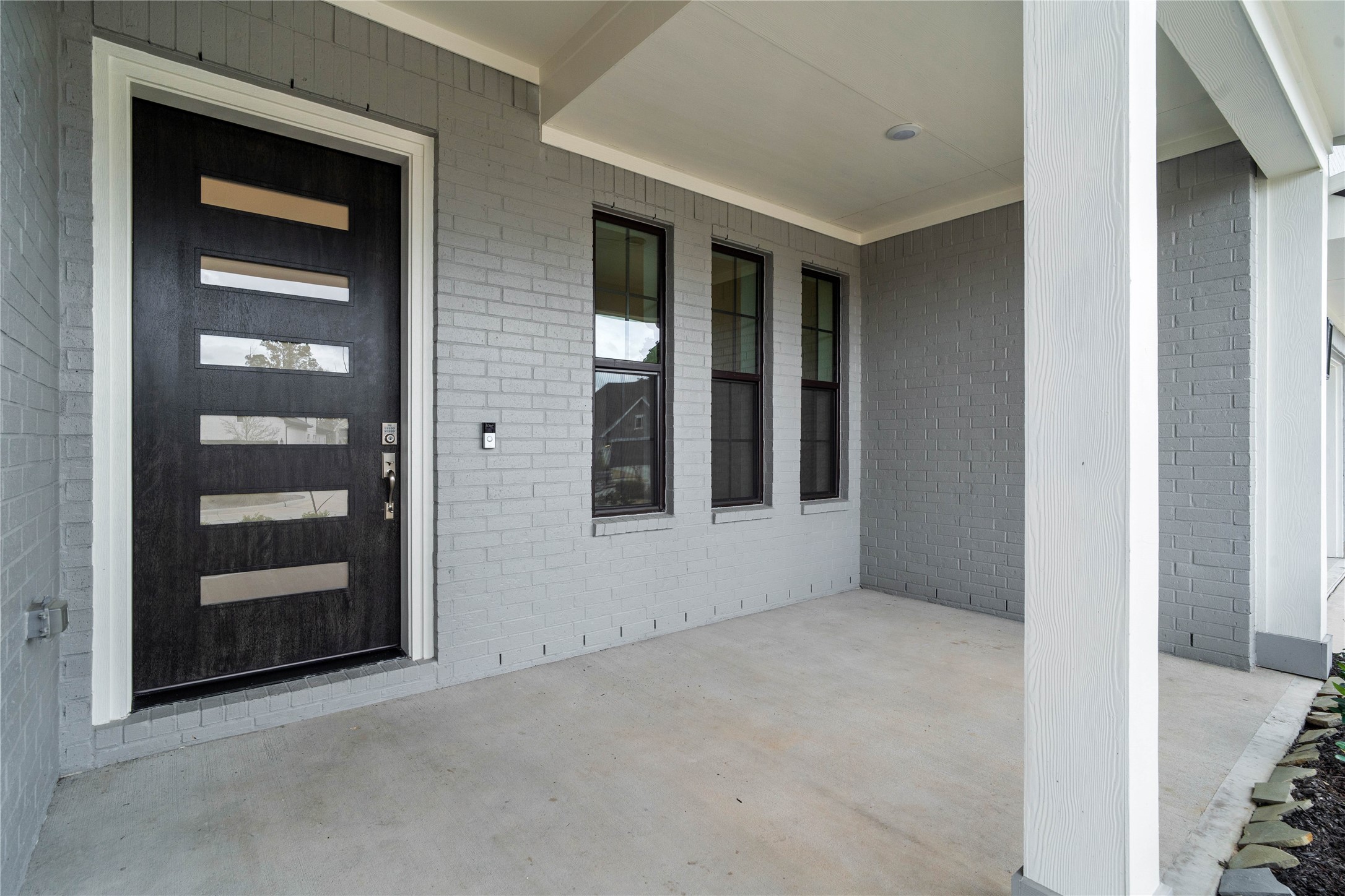 This covered front porch offers a clean, contemporary feel with its sleek grey brick faade and smooth concrete flooring. The space is anchored by a stunning modern five-lite glass front door in a rich dark finish, which provides an inviting and sophisticated entrance to the home. This covered front porch offers a clean, contemporary feel with its sleek grey brick faade and smooth concrete flooring. The space is anchored by a stunning modern five-lite glass front door in a rich dark finish, which provides an inviting and sophisticated entrance to the home.