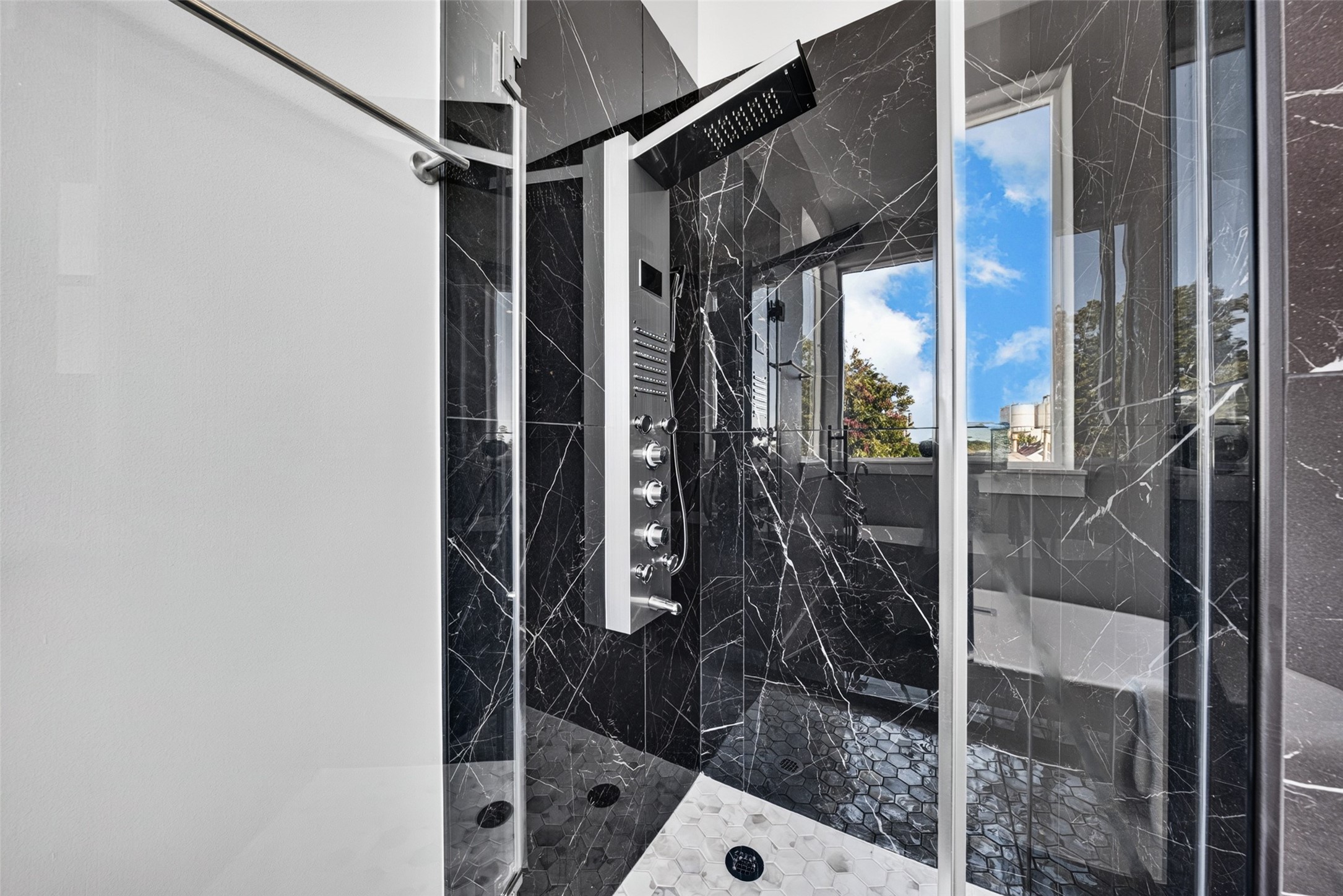 Luxurious primary walk-in shower featuring a multi-function rainfall shower panel, dramatic dark marble wall tile, glass enclosure, and modern hexagon floor tile.